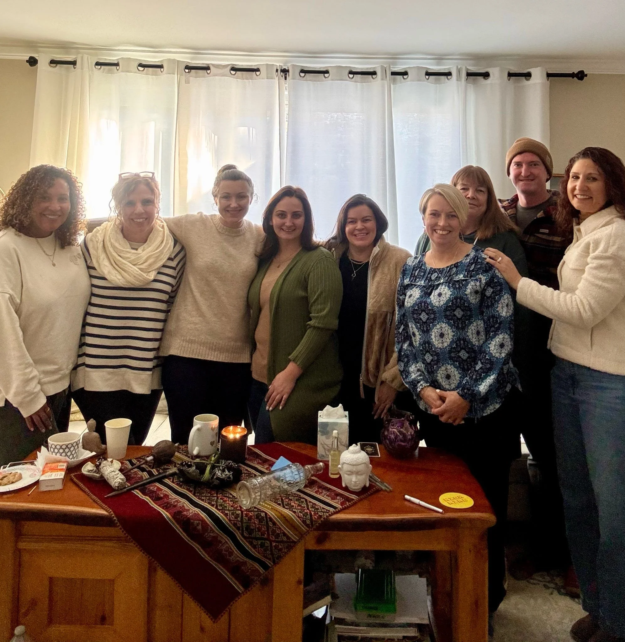 A group of ten people standing together in a living room, smiling for a photo. There is a table in front of them with various objects, including candles, cups, and decorative items.