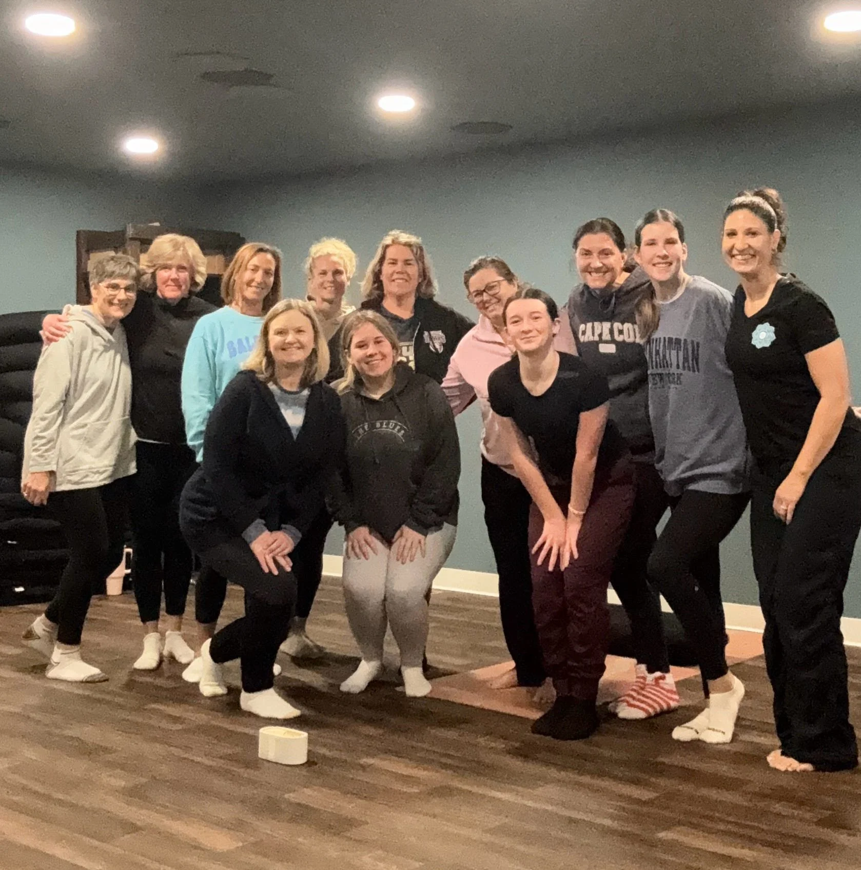 Group of women and girls posing together in a room with blue walls and wood floors, some are standing and some are kneeling, smiling at the camera.