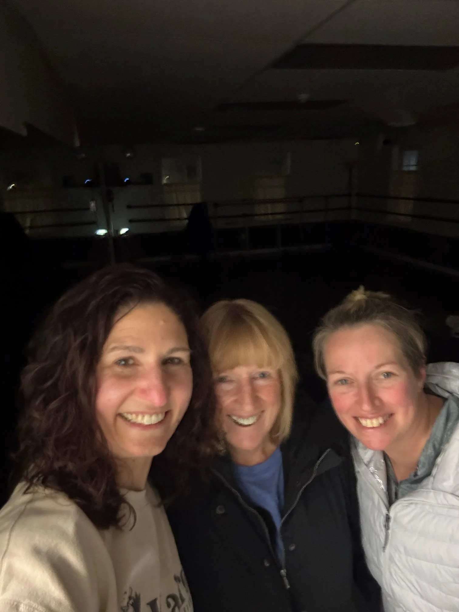Three women smiling together indoors in a dimly lit gym or dance studio.