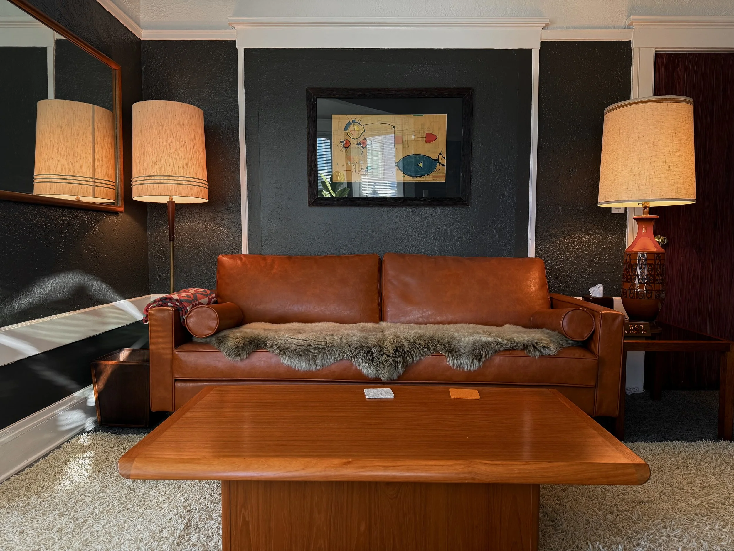 Living room with black textured walls, framed abstract art, two beige lamps on side tables, a leather sofa with a furry throw, hardwood coffee table, and a digital clock showing 6:57.