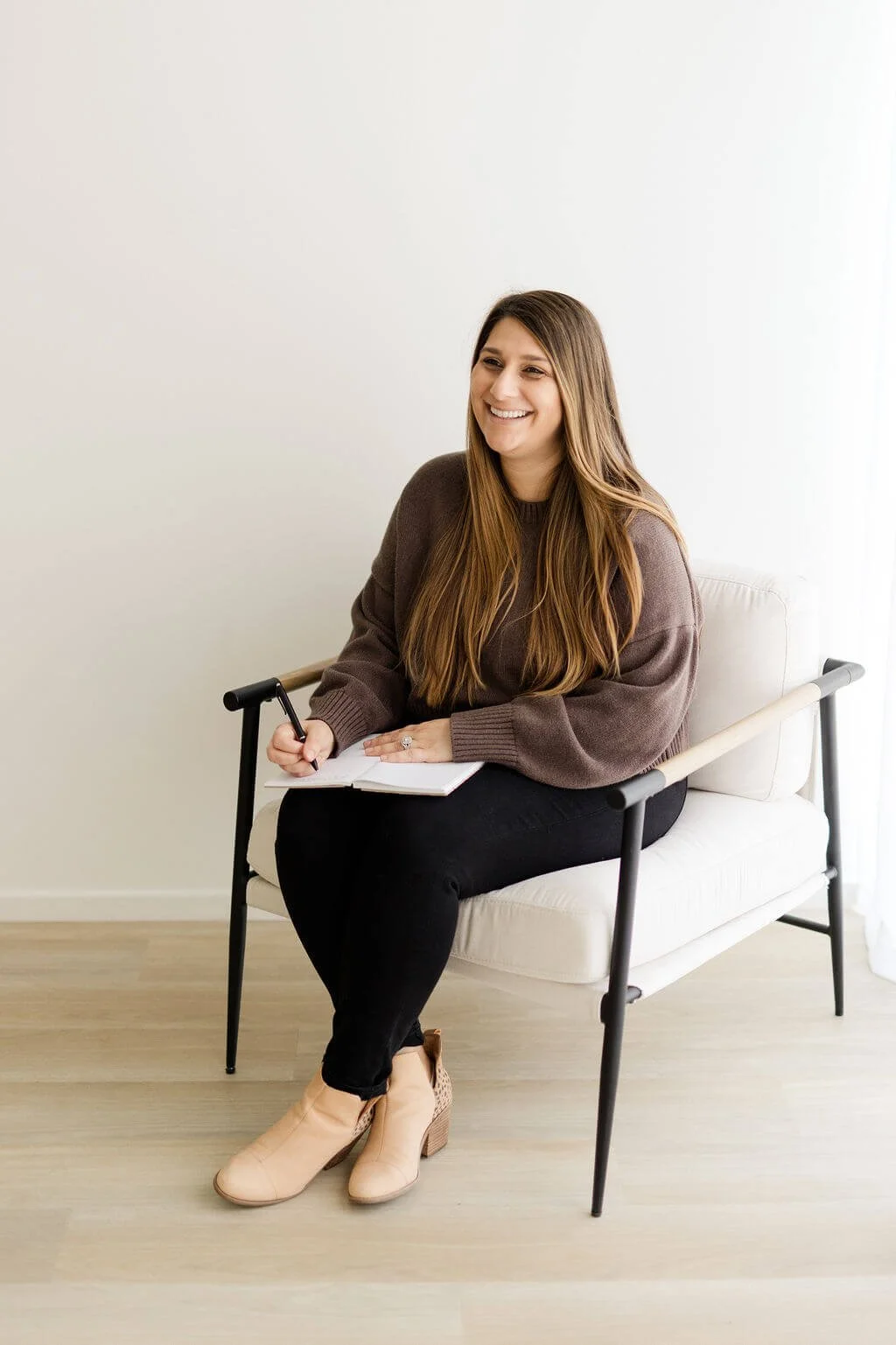oc bipoc therapists woman brown hair brown sweater black pants sitting in chair smiling writing in notebook