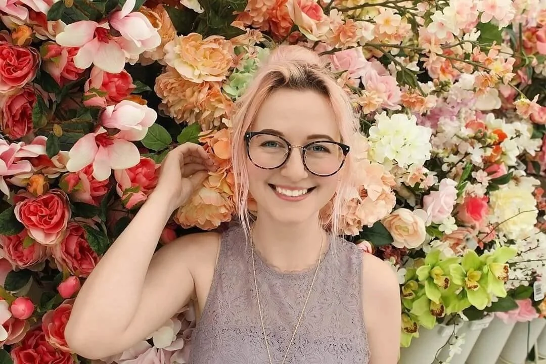 oc brainspotting therapy woman with pink blonde hair wearing glasses smiling wearing purple-grey lace top in front of flowers