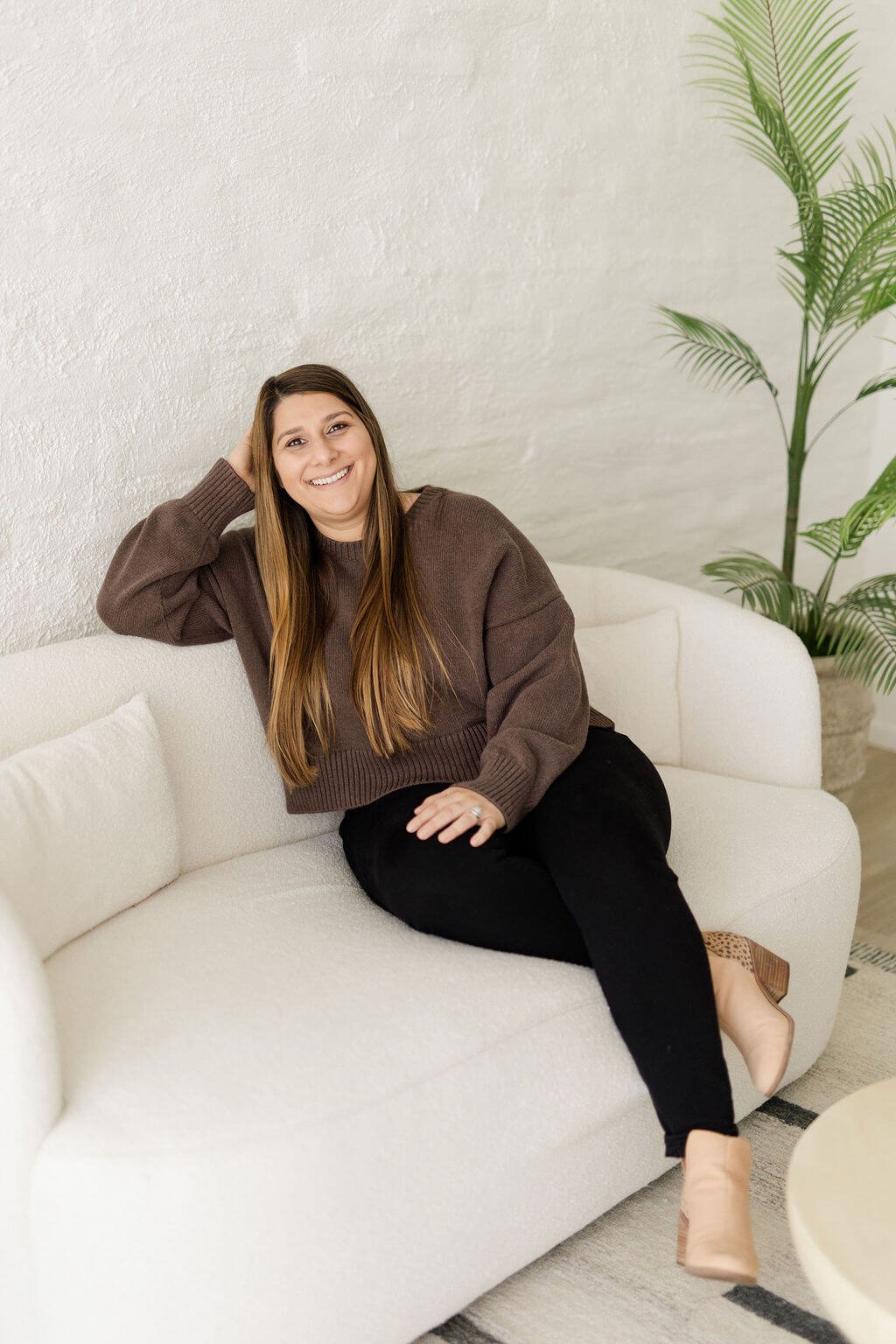 counseling in orange county woman brown hair brown top right hand resting back of head smiling sitting on white couch ankles crossed green tree in background
