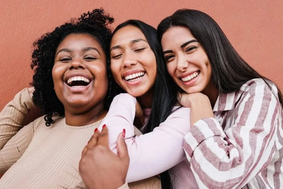 eating disorder therapist orange county 3 women of color smiling with burnt orange background wearing tan peach and striped long sleeve shirts woman on left red acrylic nails