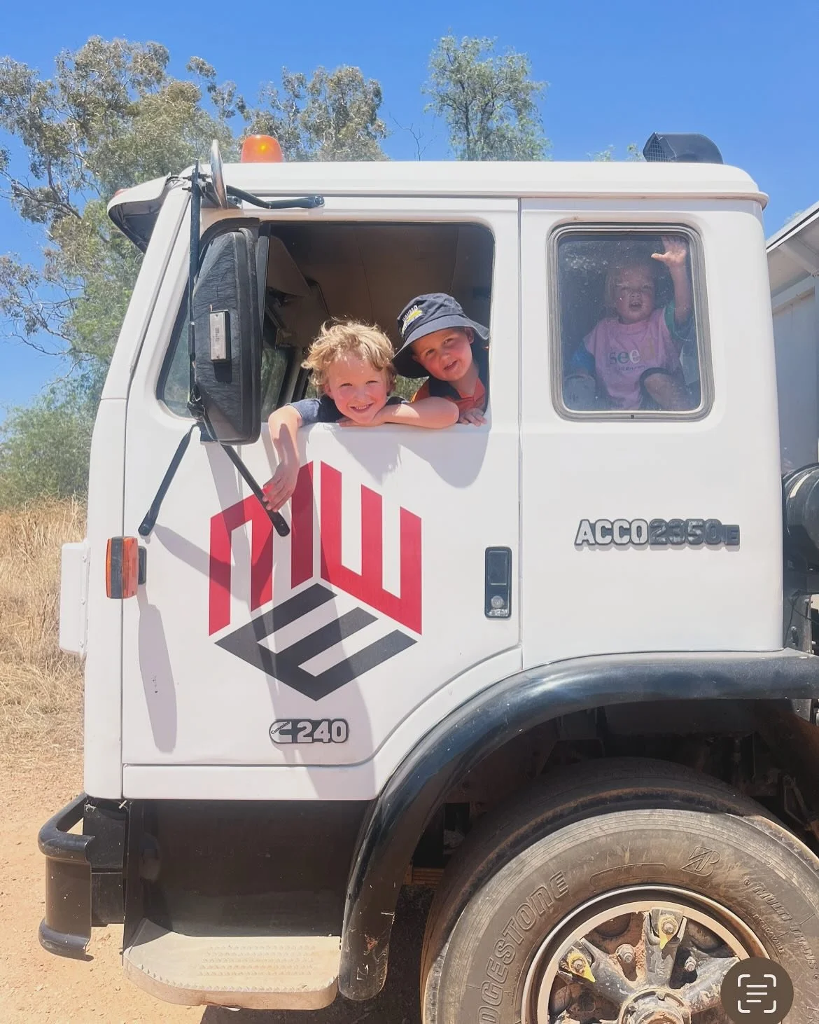 Three children smiling and leaning out of the windows of a white utility truck, with two children in the driver's cab and one in the passenger's side window, on a sunny day outdoors.
