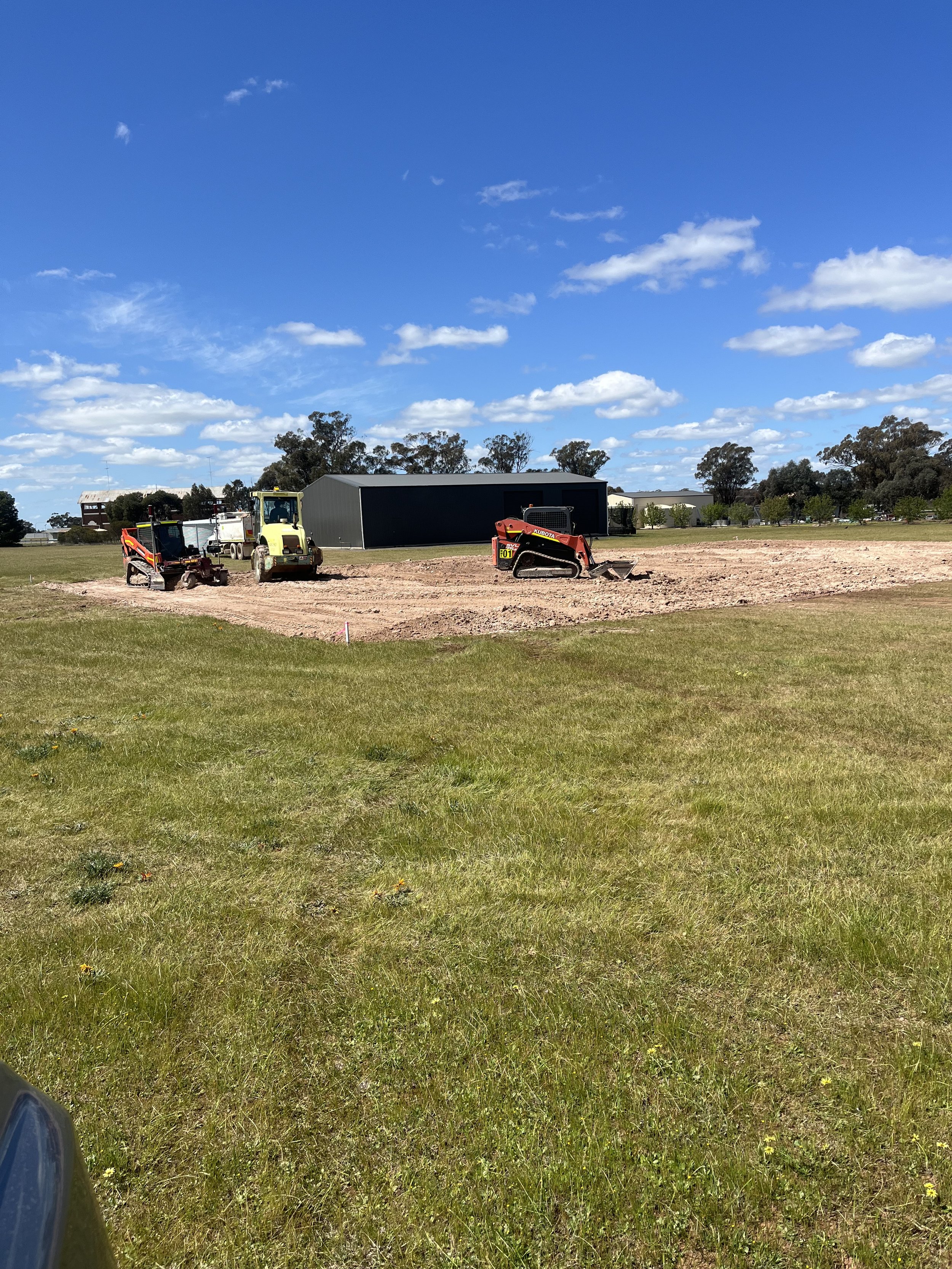 Construction site with machinery on dirt field, large gray building in background, trees, and a partly cloudy blue sky.