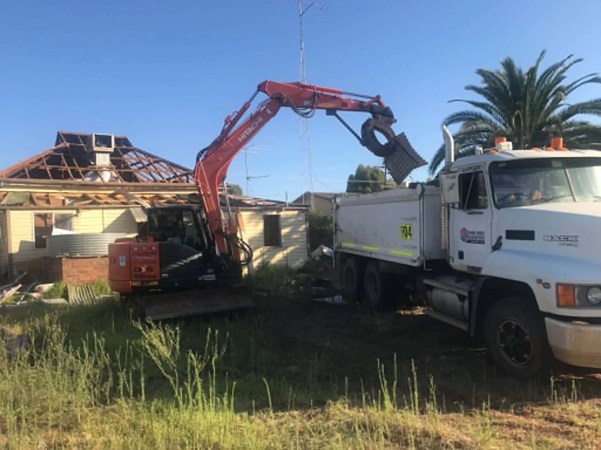 A demolistion with a truck and a hydraulic excavator demolishing a house roof with tiling. The sky is blue, and there's a palm tree in the background.