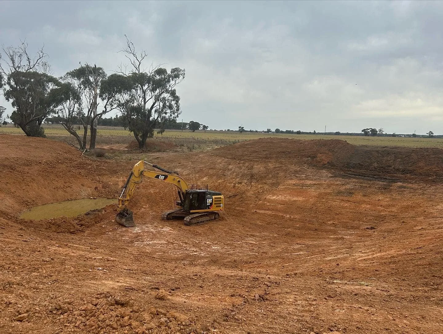 A yellow Caterpillar excavator digging in a large, open dirt area with trees in the background under cloudy skies.