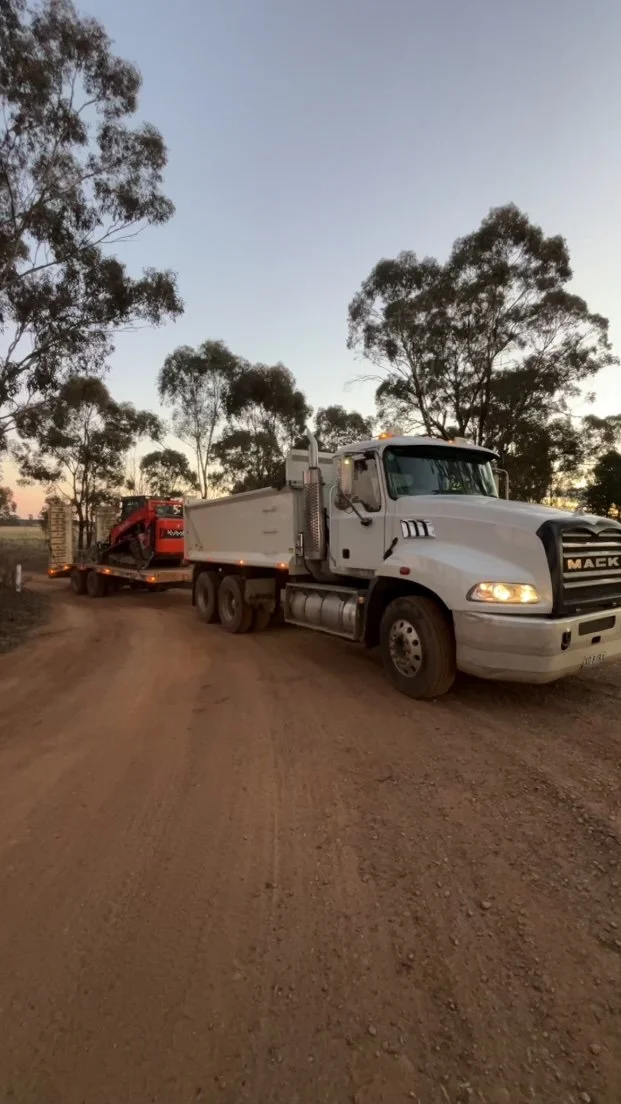 A white Mack truck carrying a red piece of construction equipment, parked on a dirt road with trees in the background during dusk.
