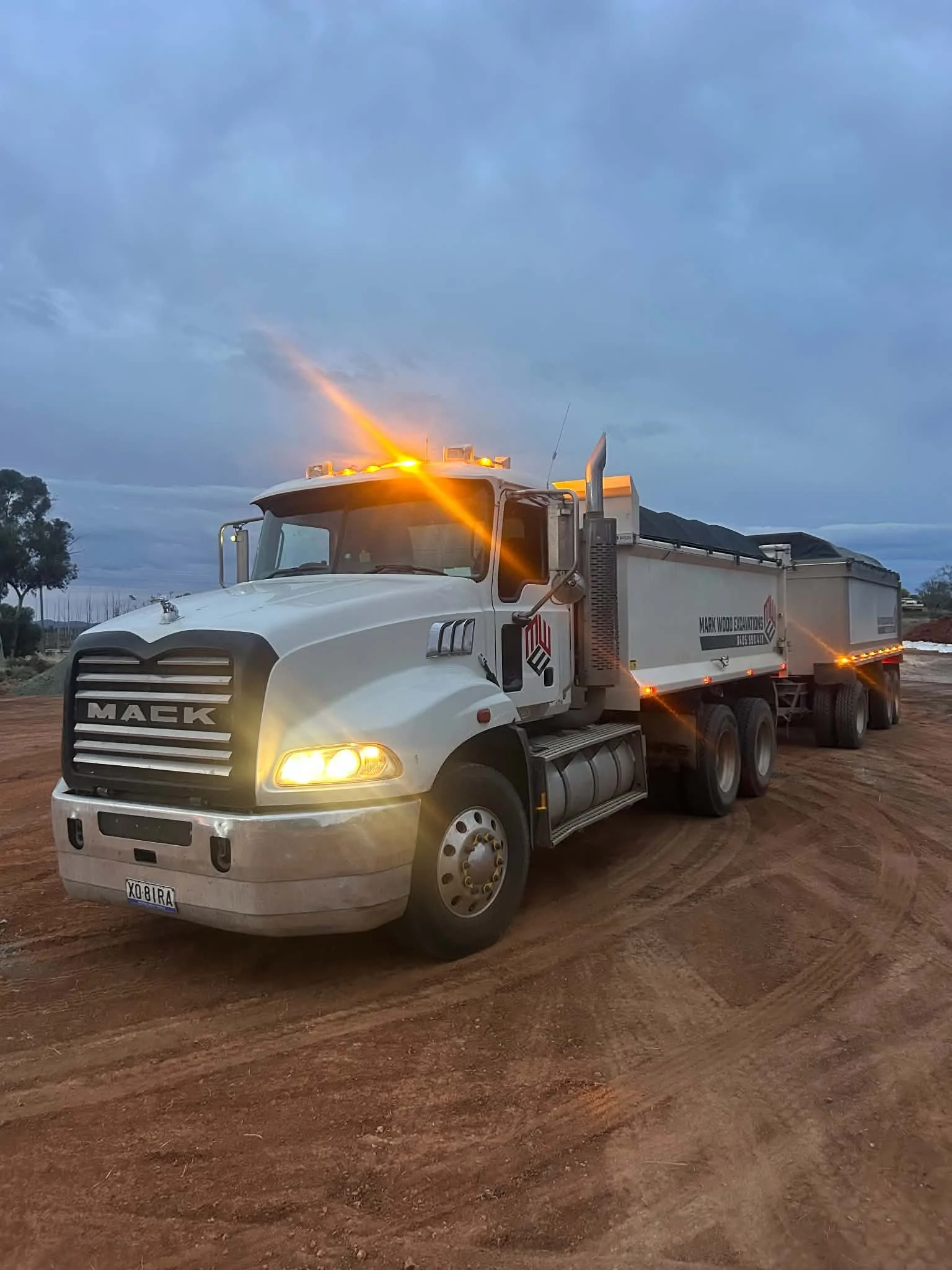 A white Mack truck with orange lights on the roof is parked on a dirt surface. The sky is partly cloudy with some blue visible.
