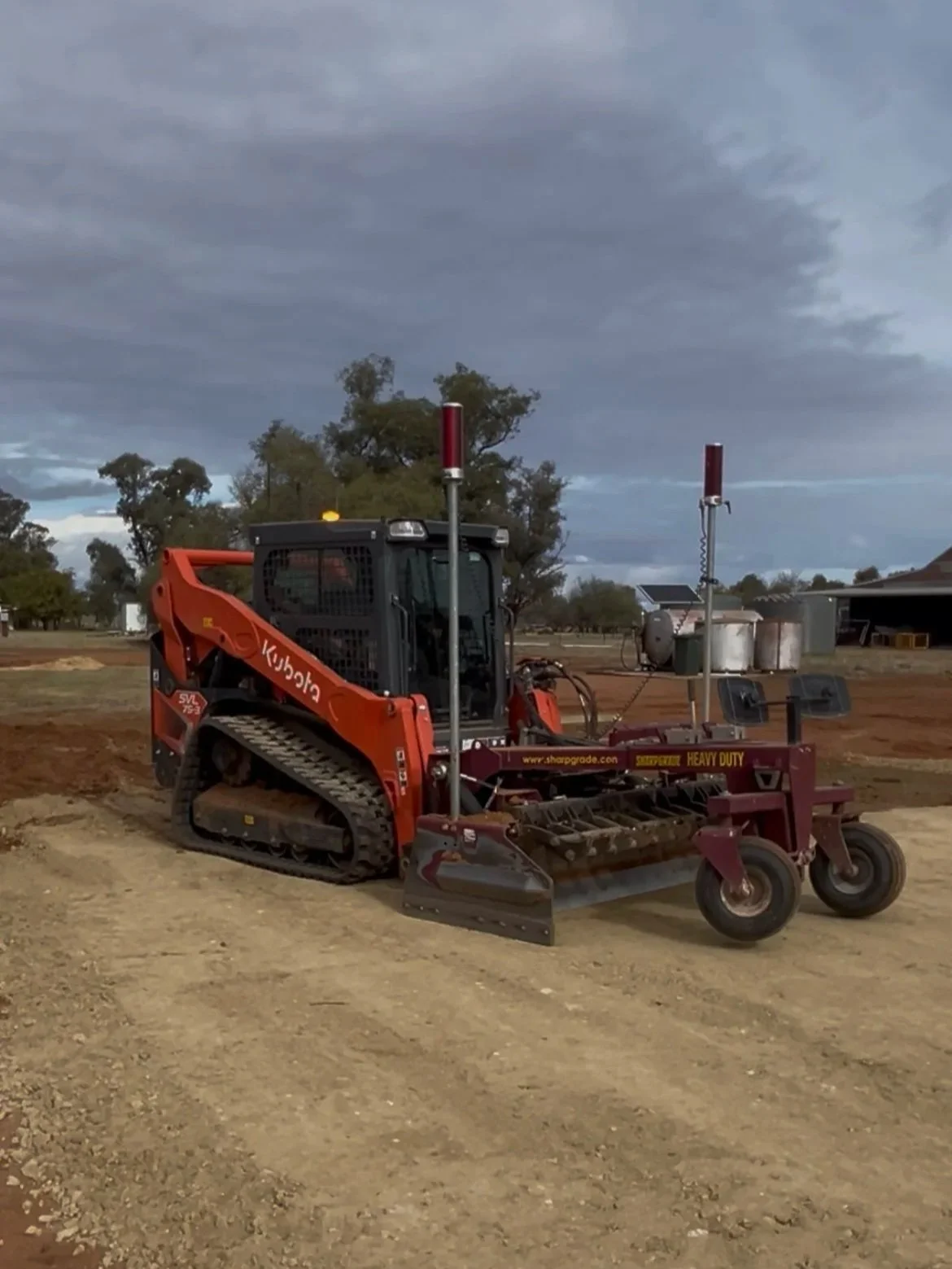 A bobcat specifically a Kubota compact track loader, equipped with a heavy-duty mower attachment on a dirt lot under a cloudy sky. The vehicle has antennas or sensors mounted on two poles.