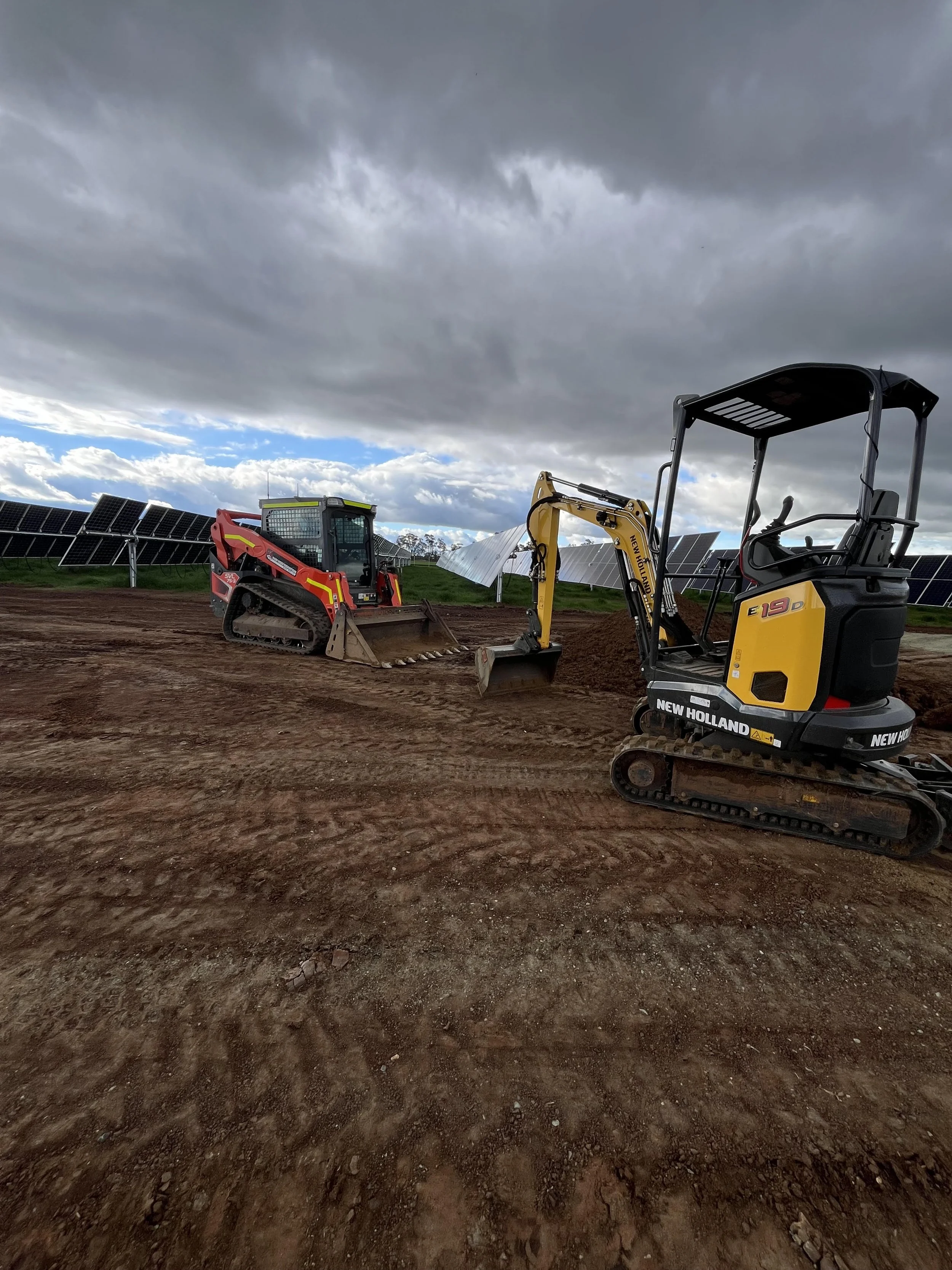 Construction site with two excavators on cleared land, solar panels in the background, and a cloudy sky overhead.
