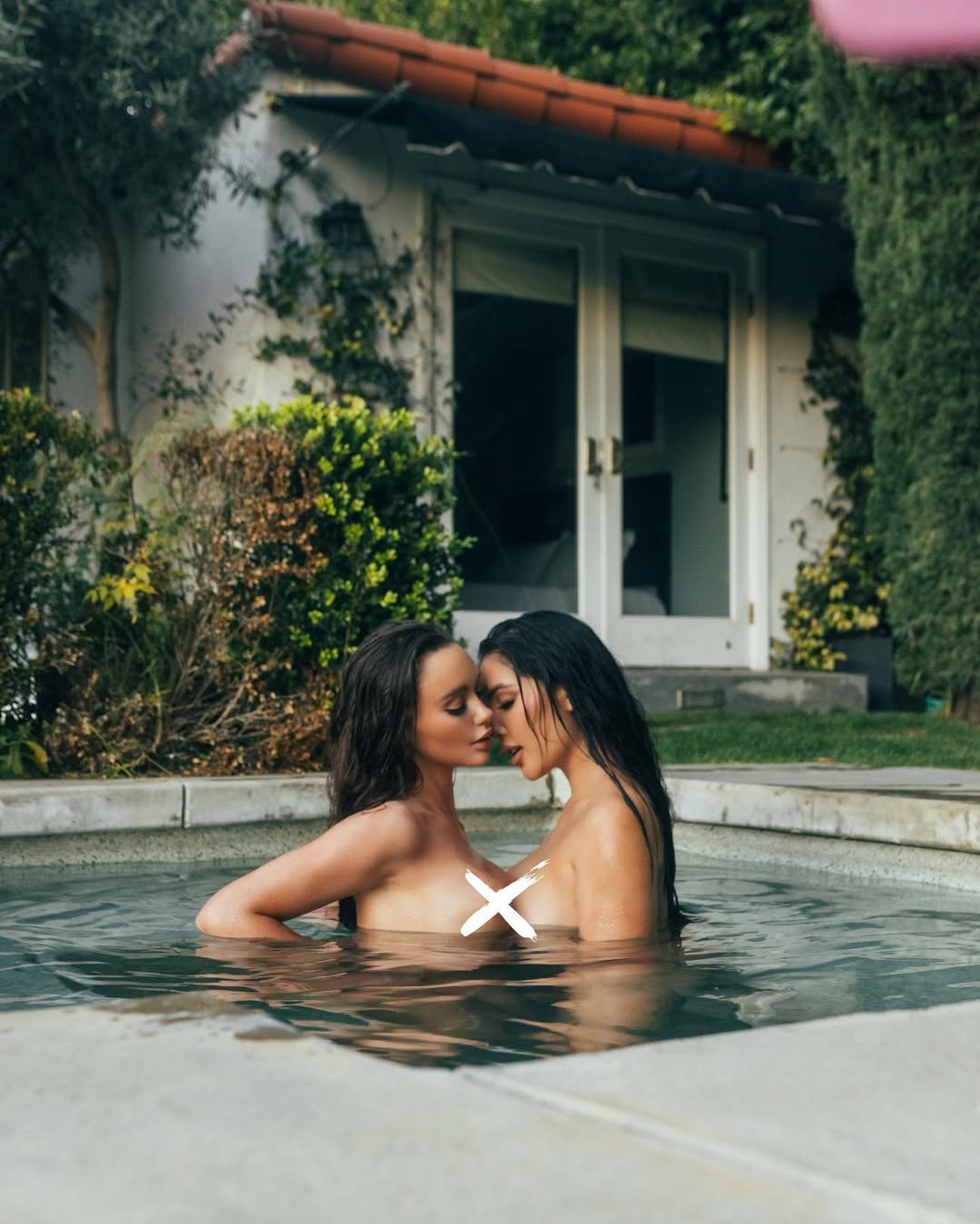 Two women with wet hair and no swimsuits embracing in a hot tub outdoors, with a house and greenery in the background.