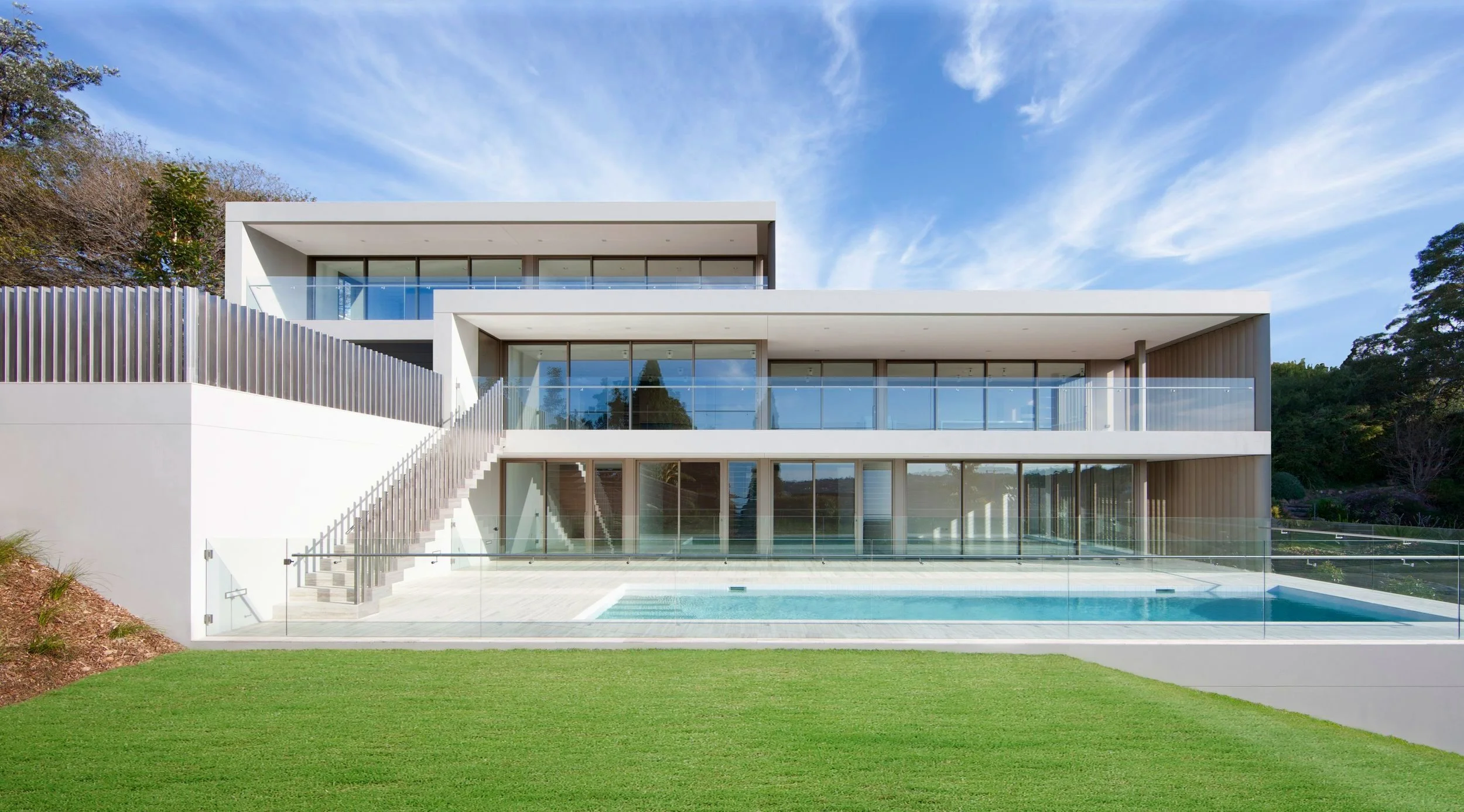 Modern three-story house with large glass windows, balconies with glass railings, and a swimming pool in the backyard, surrounded by green grass and trees under a blue sky with clouds.