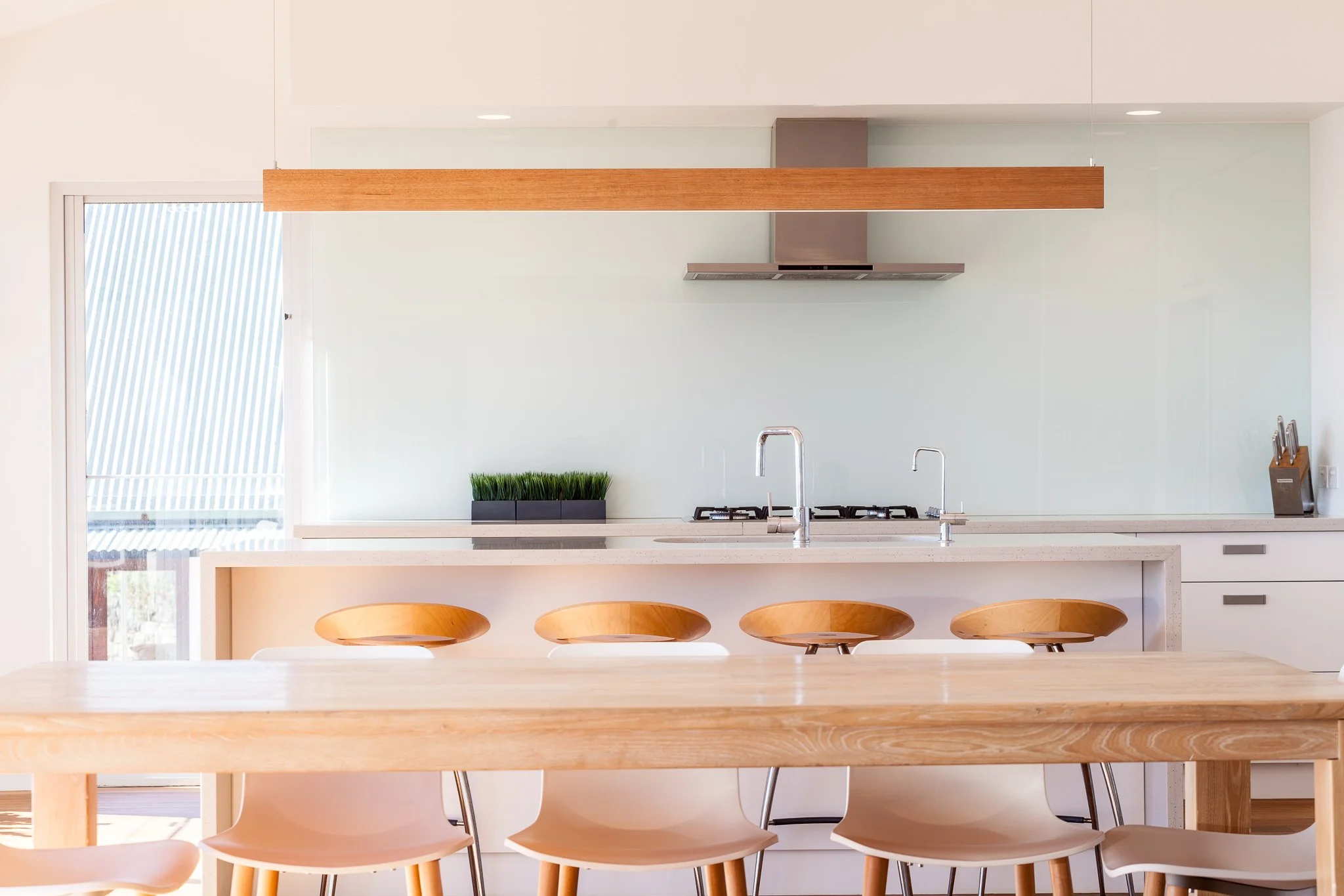 A modern kitchen with light-colored wooden dining table and chairs, white cabinetry, a countertop with a sink, a gas stove, and a green plant on the counter.