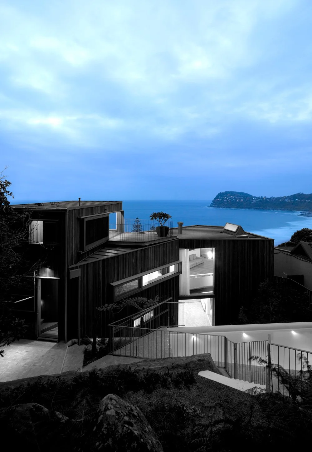 Modern house with black wooden exterior, illuminated windows, rooftop terrace with potted plant, overlooking ocean and distant hillside under cloudy sky during dusk.