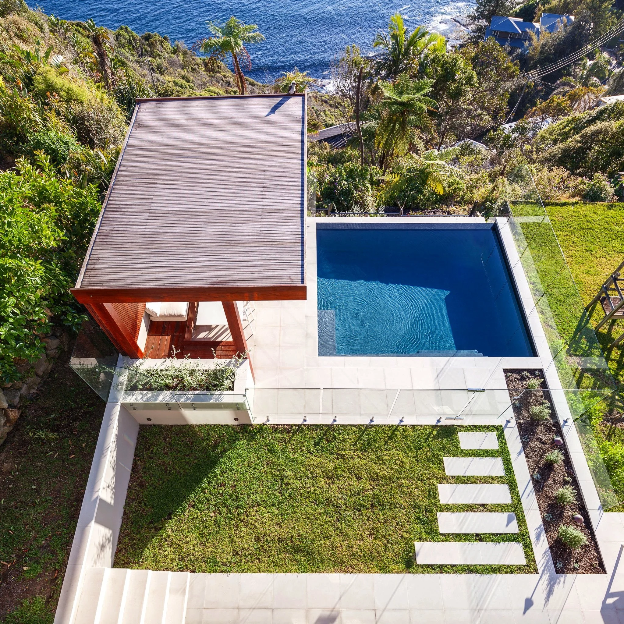 Aerial view of a modern backyard with a swimming pool, a grassy area with stepping stones, a wooden deck with a shaded seating area, and lush greenery overlooking the ocean in the background.
