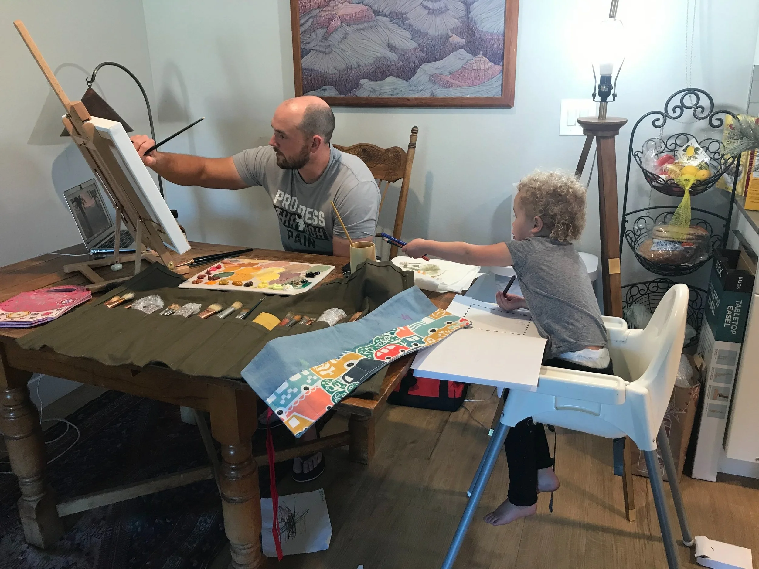 A man is painting on an easel while a young girl in a high chair reaches for his paintbrush at a dining table.