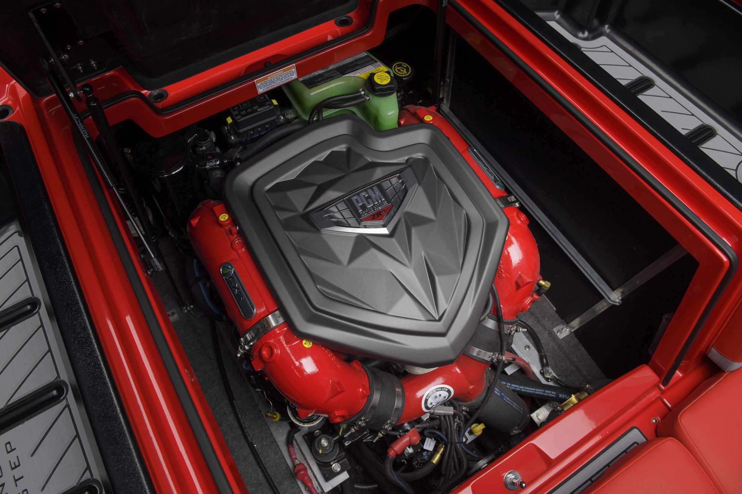 The engine bay of a red vehicle featuring a prominent supercharged engine with a large, angular gray cover labeled "PCM ZR1." The engine components are red with black and green accents, and the surrounding area includes various hoses, wires, and metallic parts, with some labels and warning stickers.