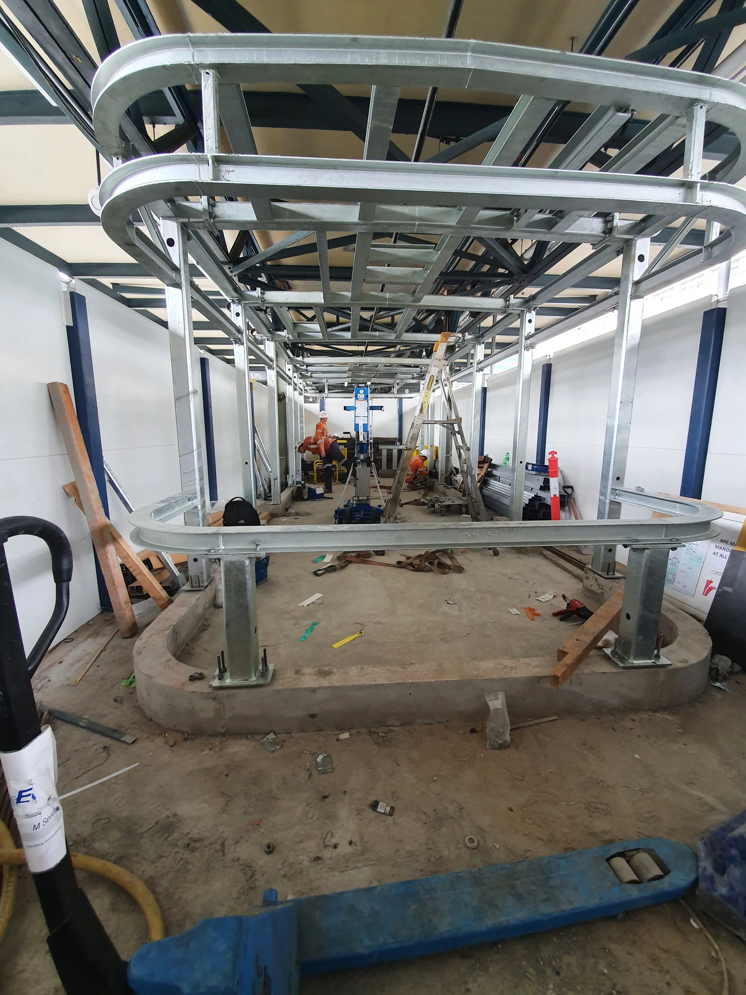 Construction site with metal framework and workers in orange safety clothing and helmets, working on a structure in an indoor space.