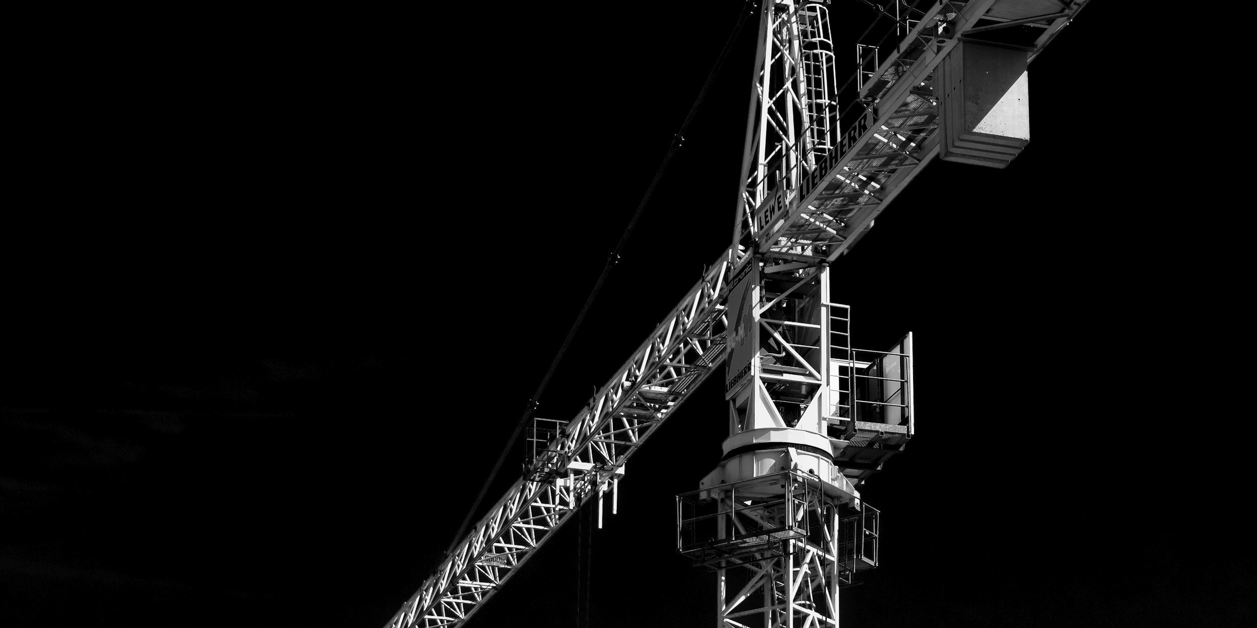 Black and white photo of a tall construction crane against a dark background, showing the lattice structure of the crane's arm and cabin.