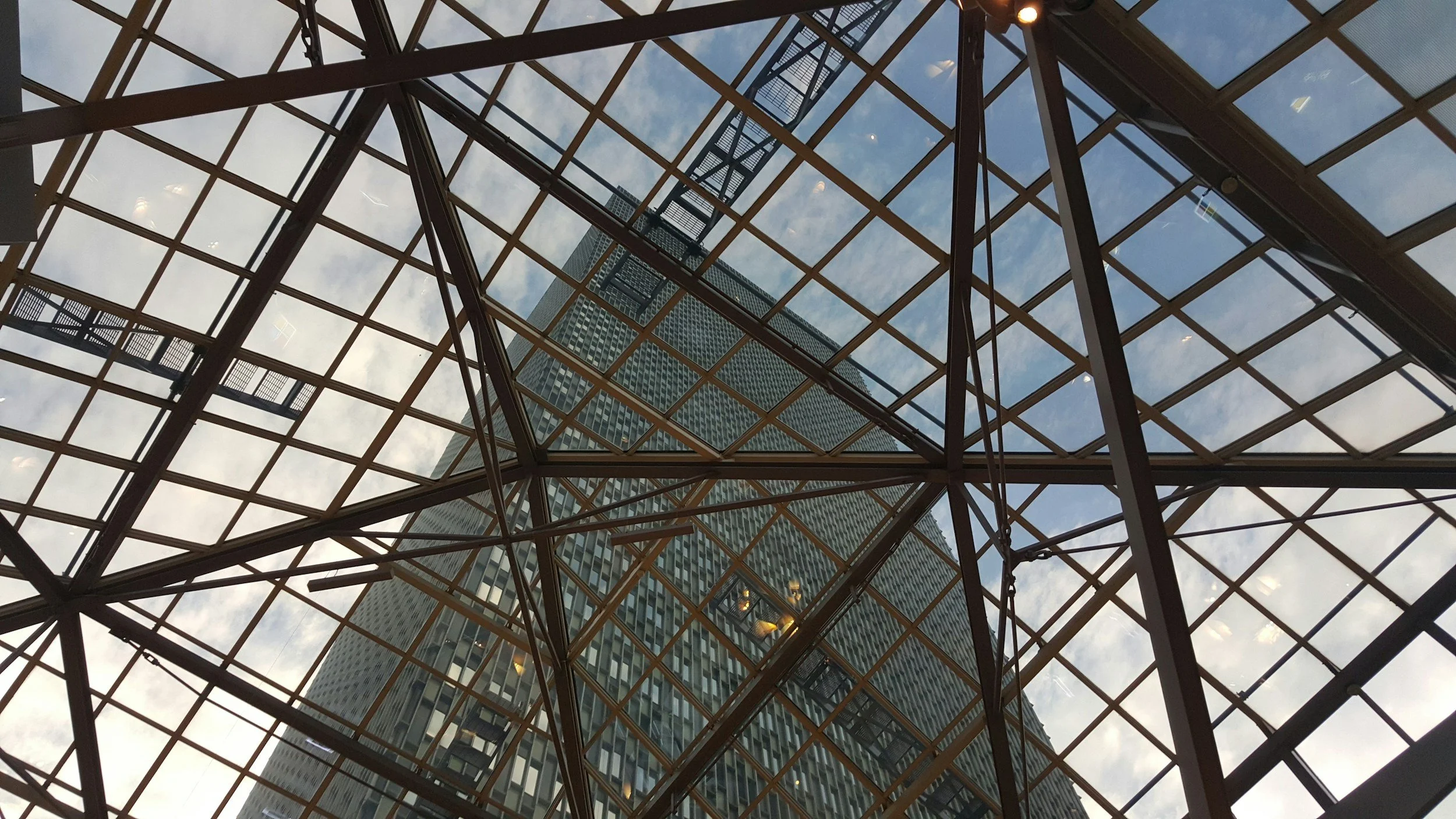 View of a modern skyscraper through the glass ceiling of a building, with metal framework and stairs visible, set against a sky with clouds.