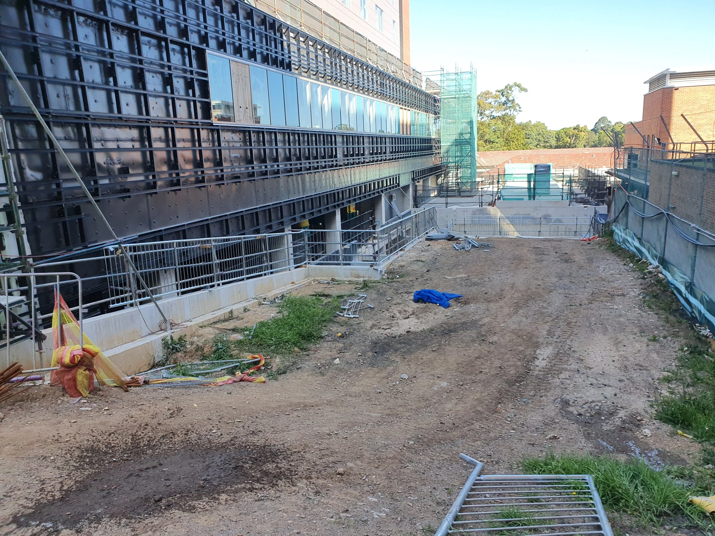 Construction site with dirt ground, metal railings, scaffolding, and partially constructed building with glass windows.