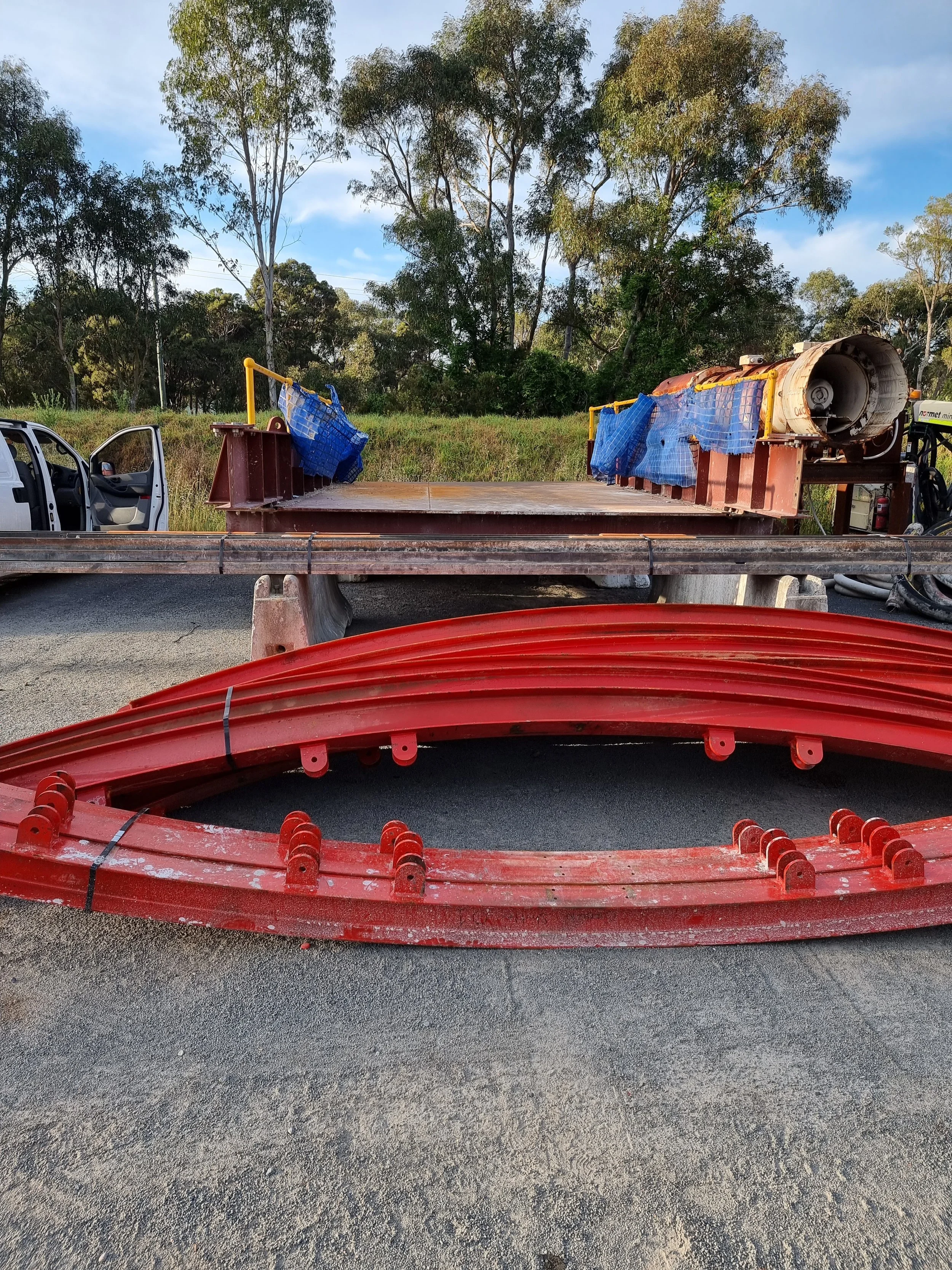 Construction site with stacked red metal beams and a flatbed truck with blue tarps covering equipment, set against trees and blue sky.