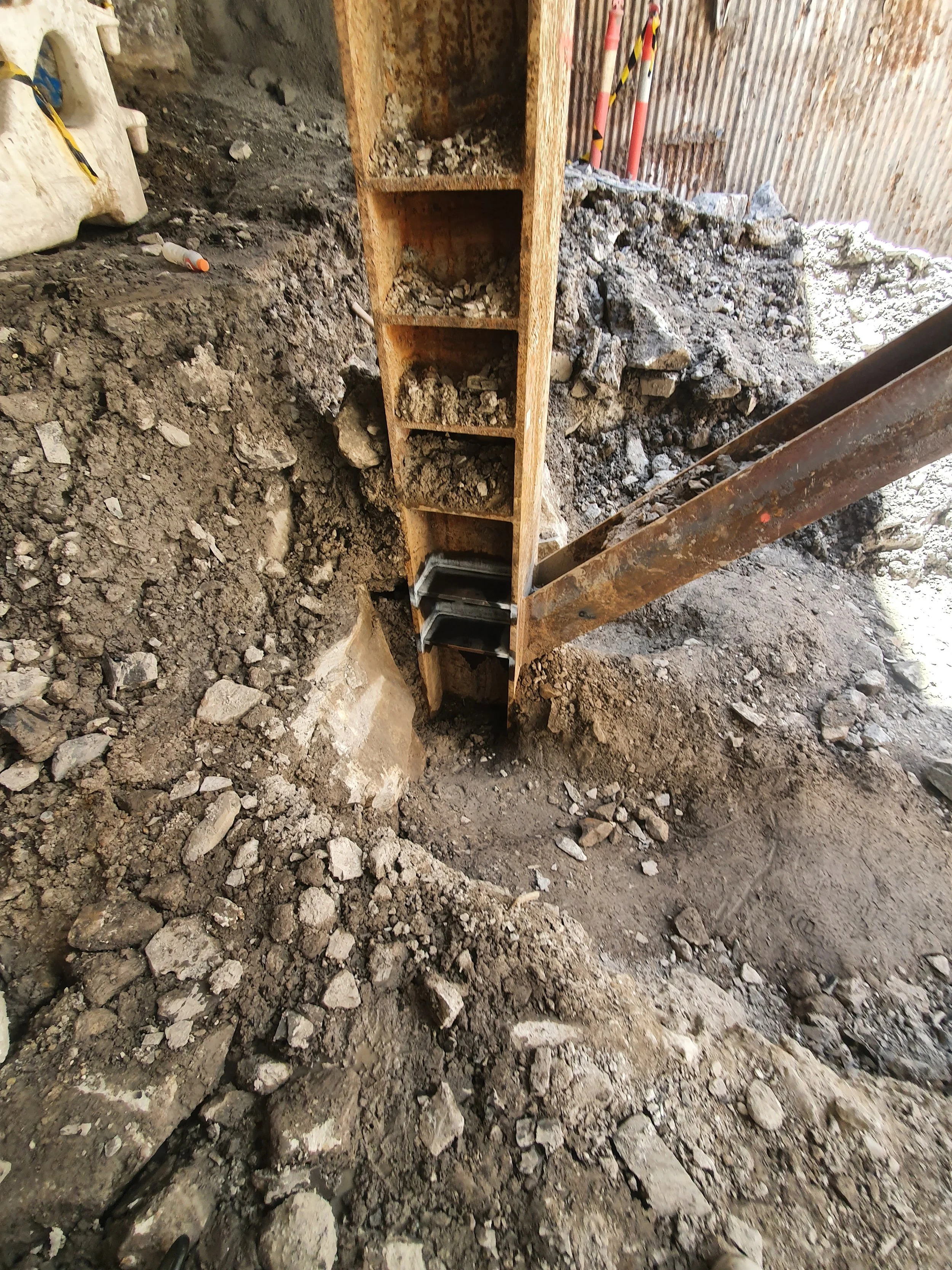 Construction site showing a vertical steel beam with a wooden support, surrounded by dirt and rubble.