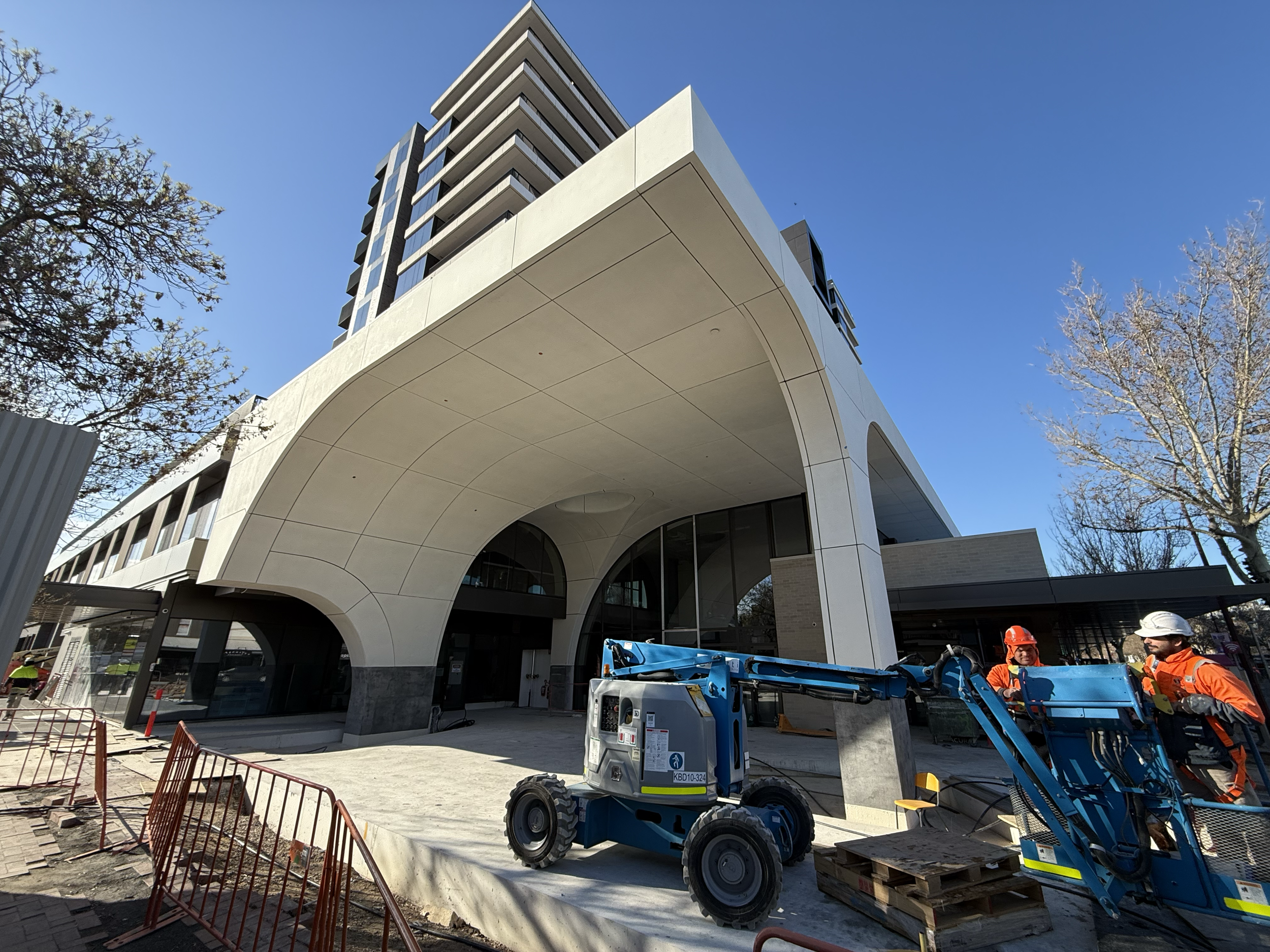 Construction workers and machinery outside a modern building with an arched entrance and tall upper floors during daytime.