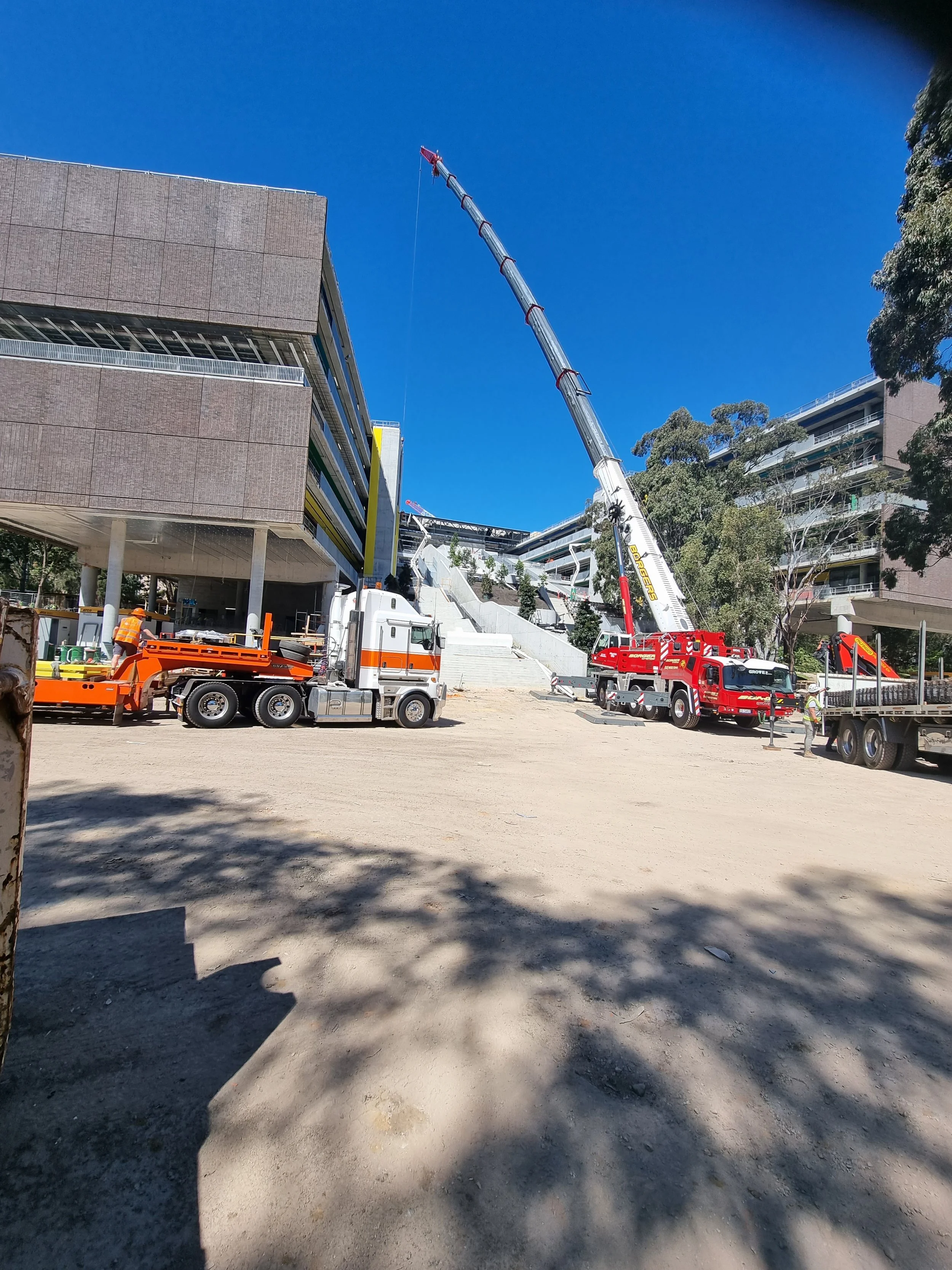Construction site with crane and trucks near modern buildings, workers in safety gear, and clear blue sky.