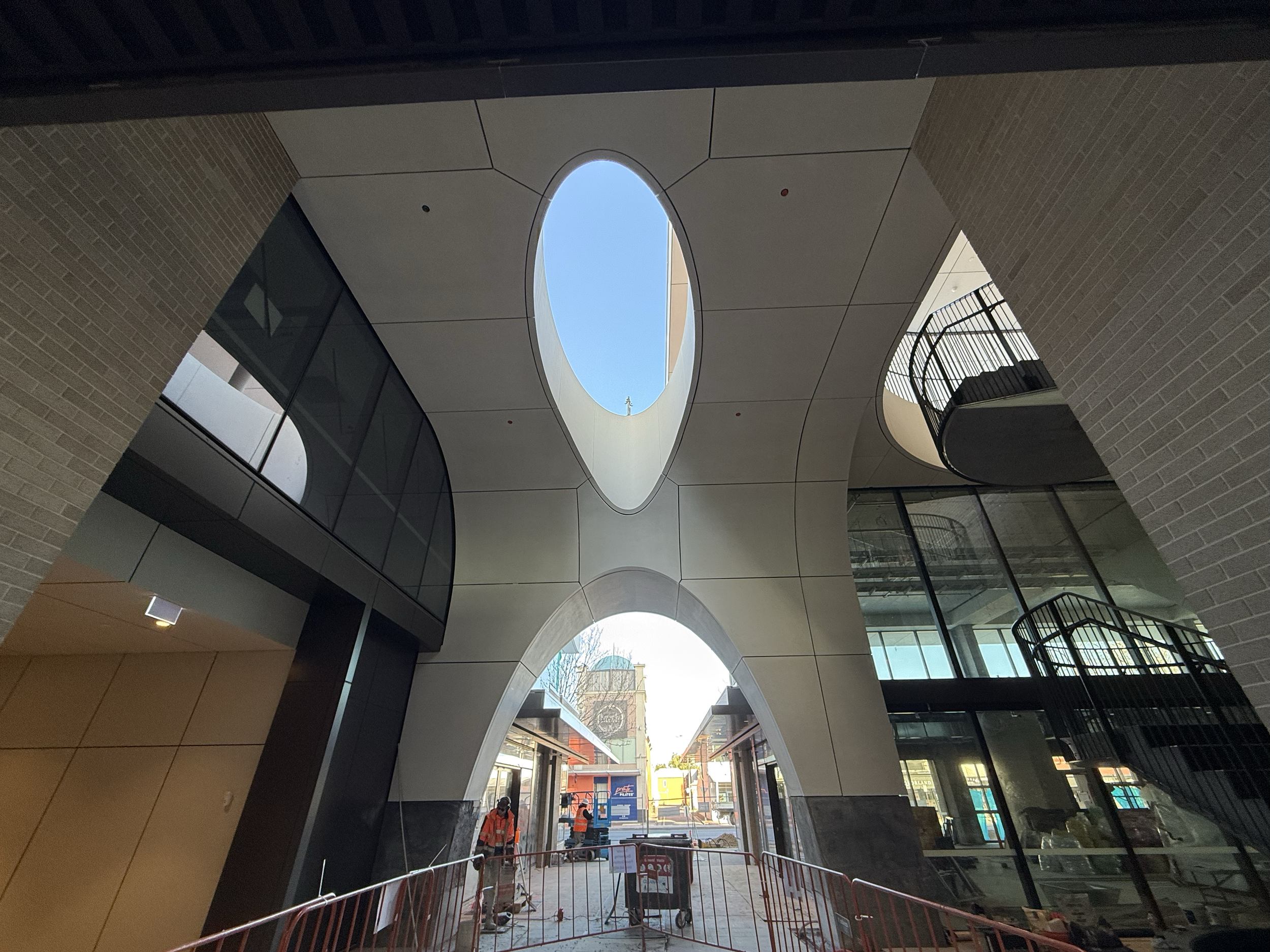 Modern building with curved architecture, large openings in the ceiling revealing a blue sky, glass walls, and a staircase. Construction workers are present outside.