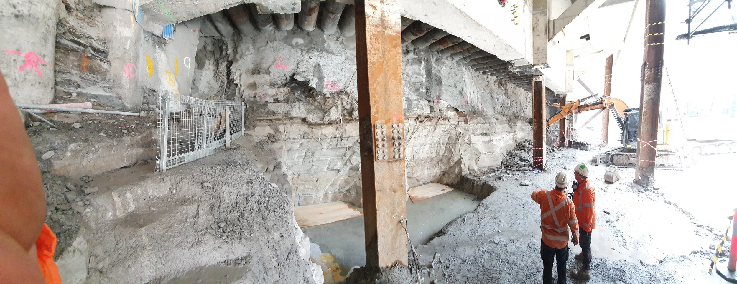 Construction workers in orange safety gear and helmets standing on a construction site with exposed dirt and large metal support beams.