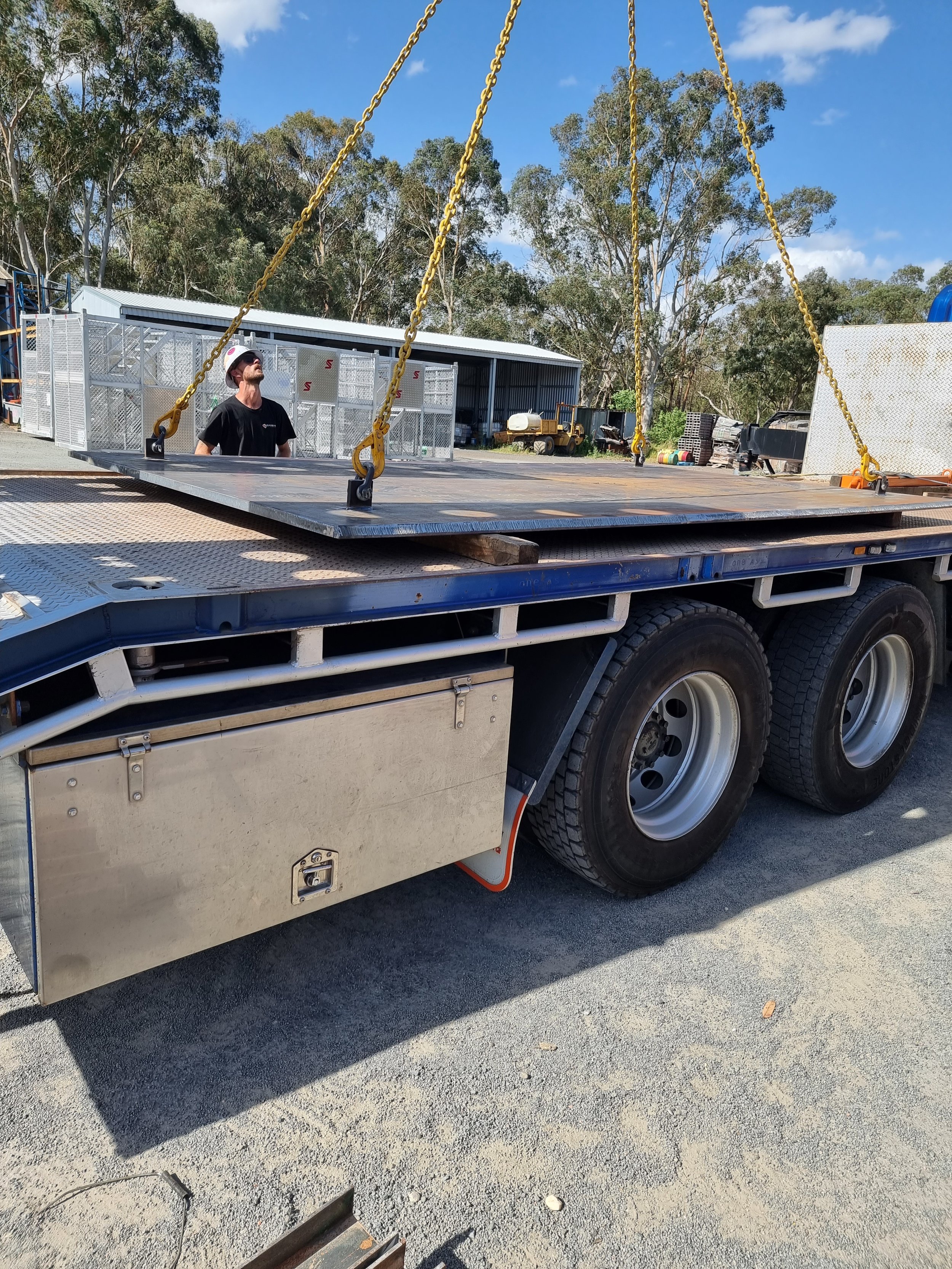 A man wearing a black shirt and white helmet standing on a flatbed truck with yellow chains hanging from its sides, under a partly cloudy blue sky with trees and industrial equipment in the background.