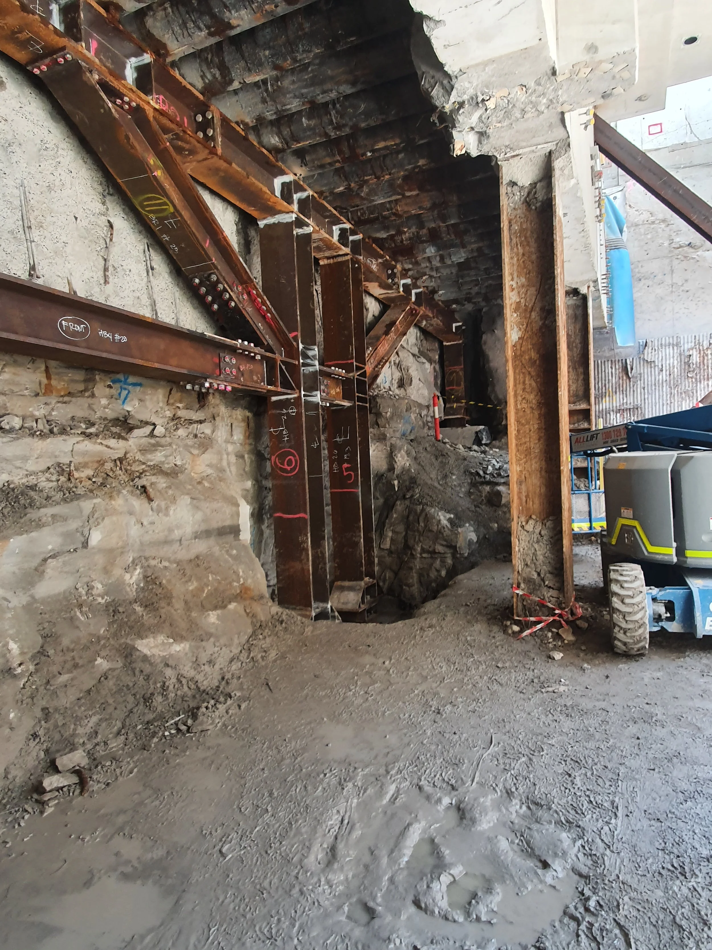 Construction site with exposed steel beams, partially demolished stone and brick wall, and construction equipment.