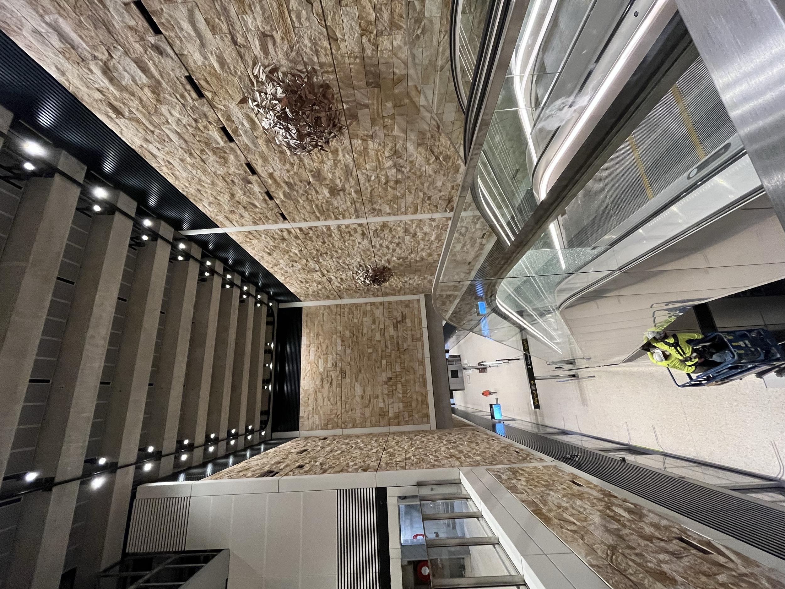 Interior of an airport terminal with stone walls, escalators, and a person in a high-visibility vest pushing a luggage cart.