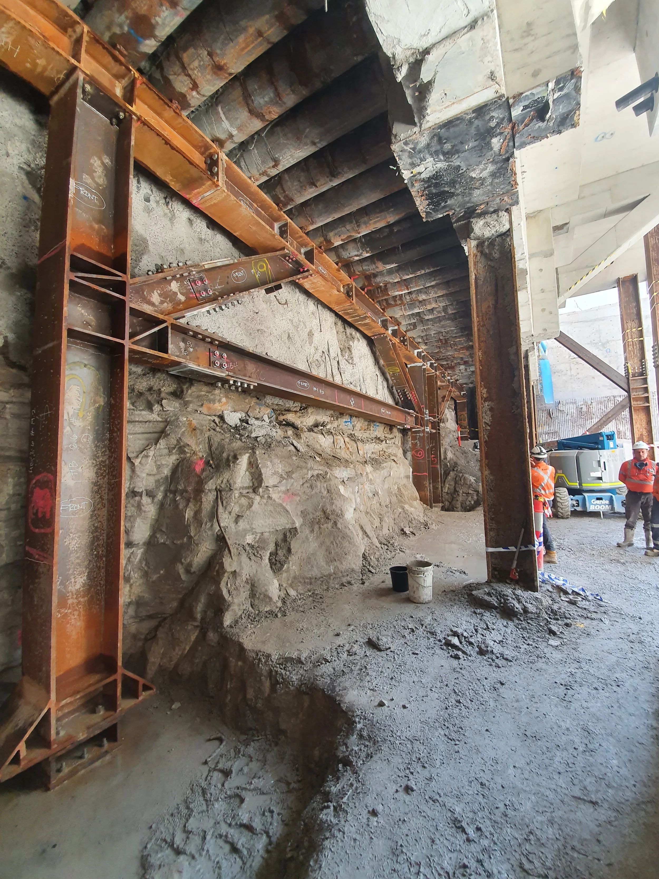 Construction site with steel supports, exposed rocks, and workers in safety gear.