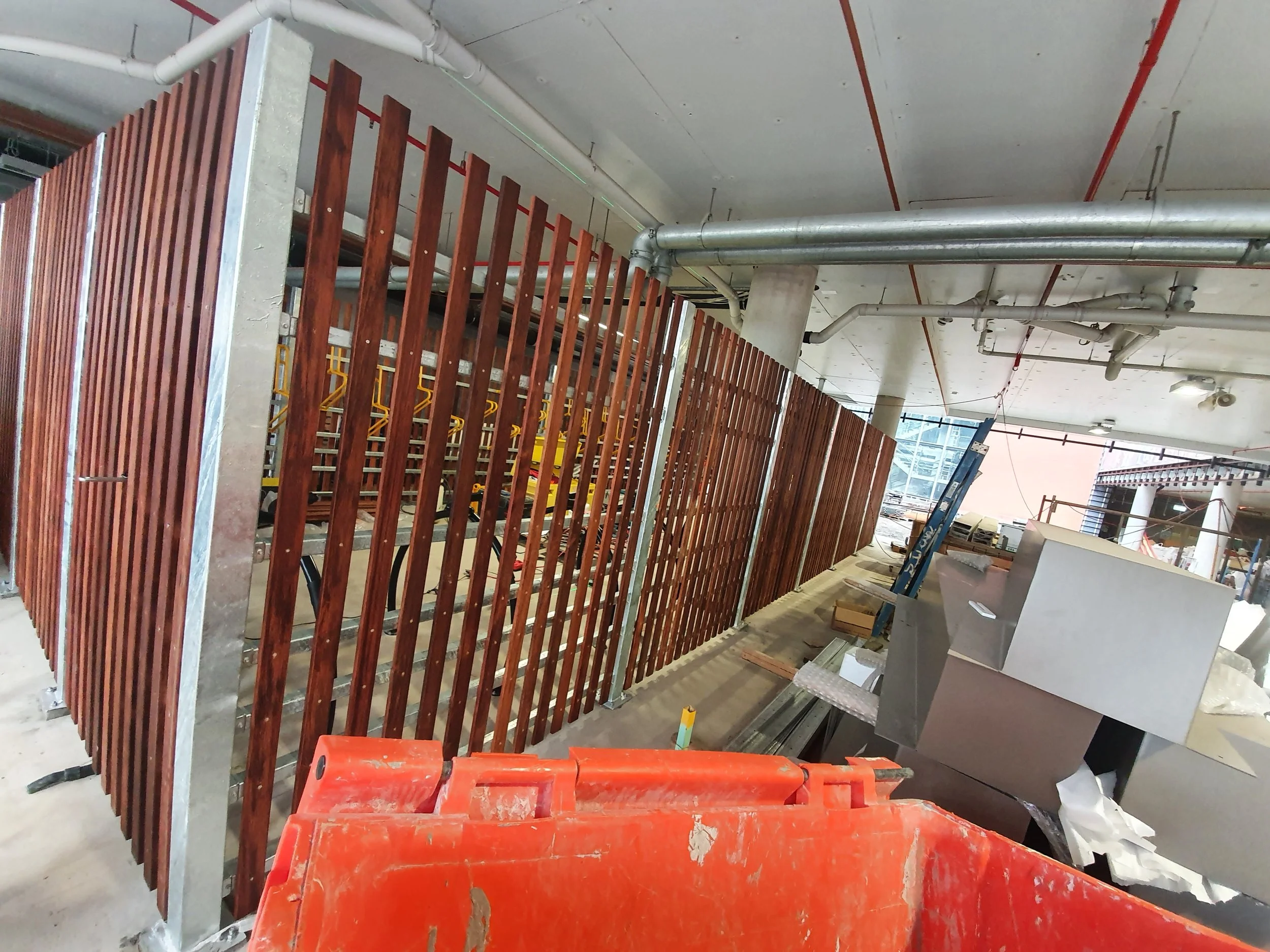 Construction site with a wooden slat fence, construction materials, and equipment, and exposed ceiling pipes.