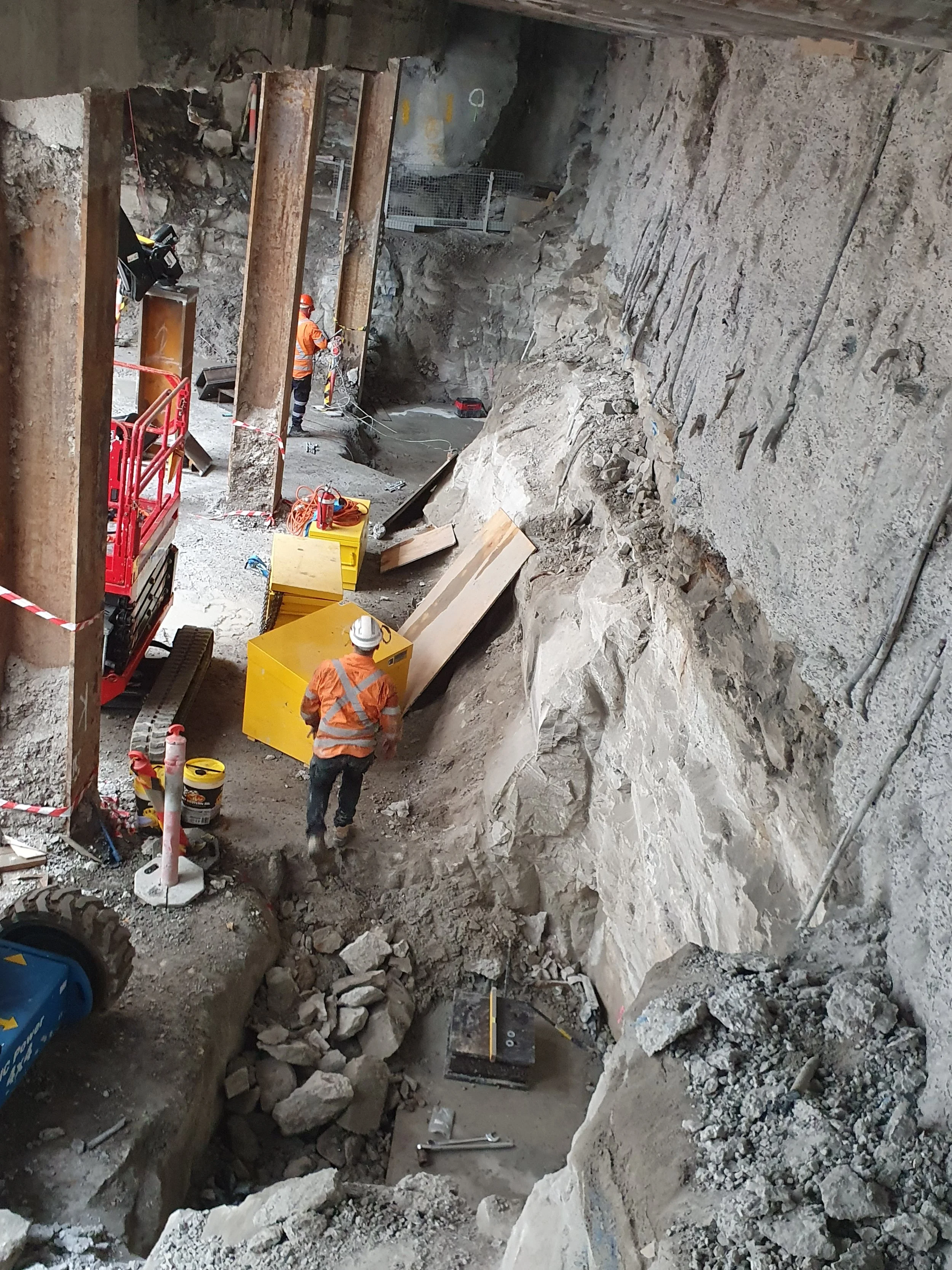 Construction workers in orange safety shirts and helmets working on a debris-filled underground construction site with large rocks, equipment, and steel supports.