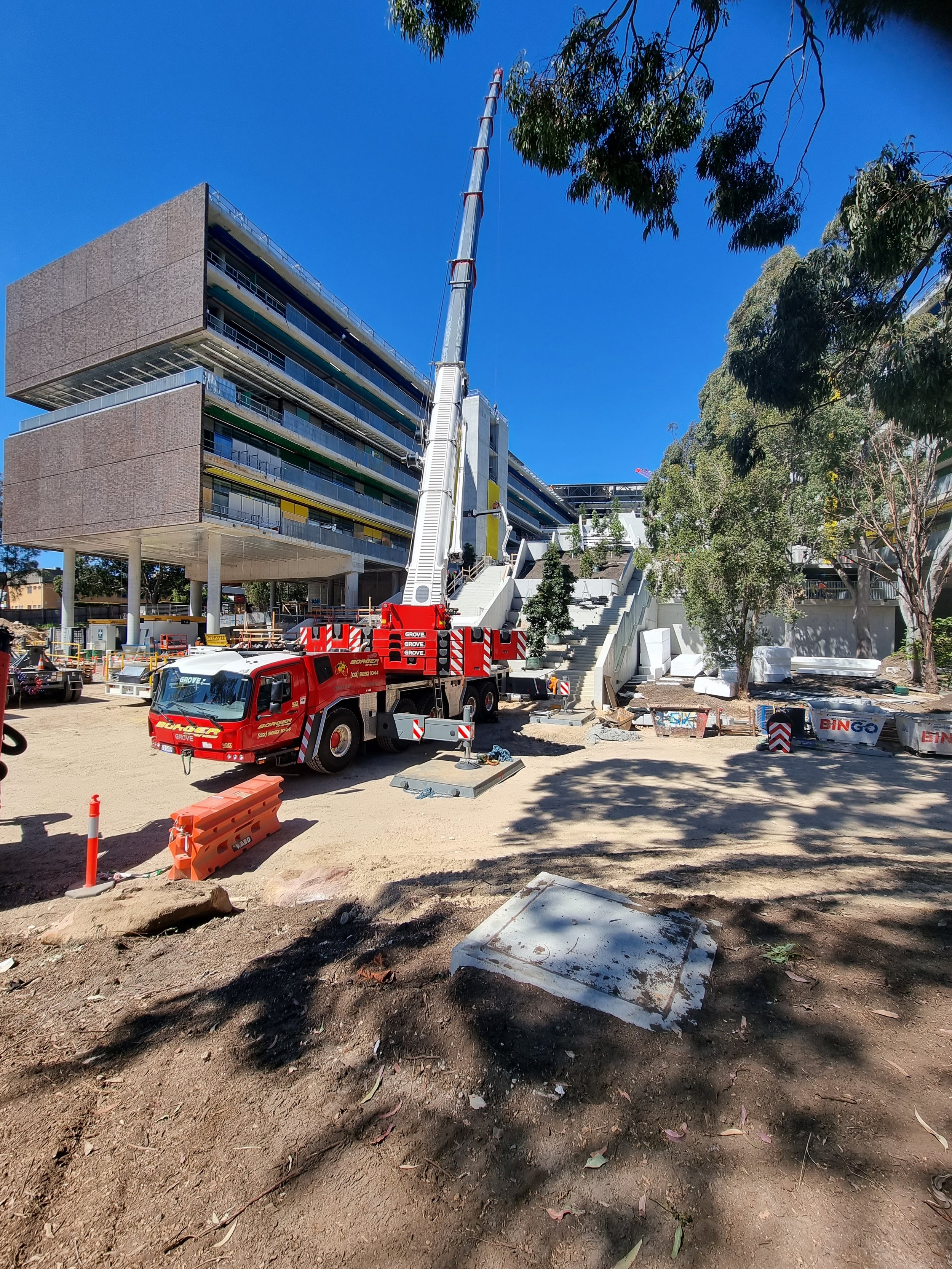 Construction site with a large crane truck in front of a modern multi-story building, surrounded by trees and construction equipment.