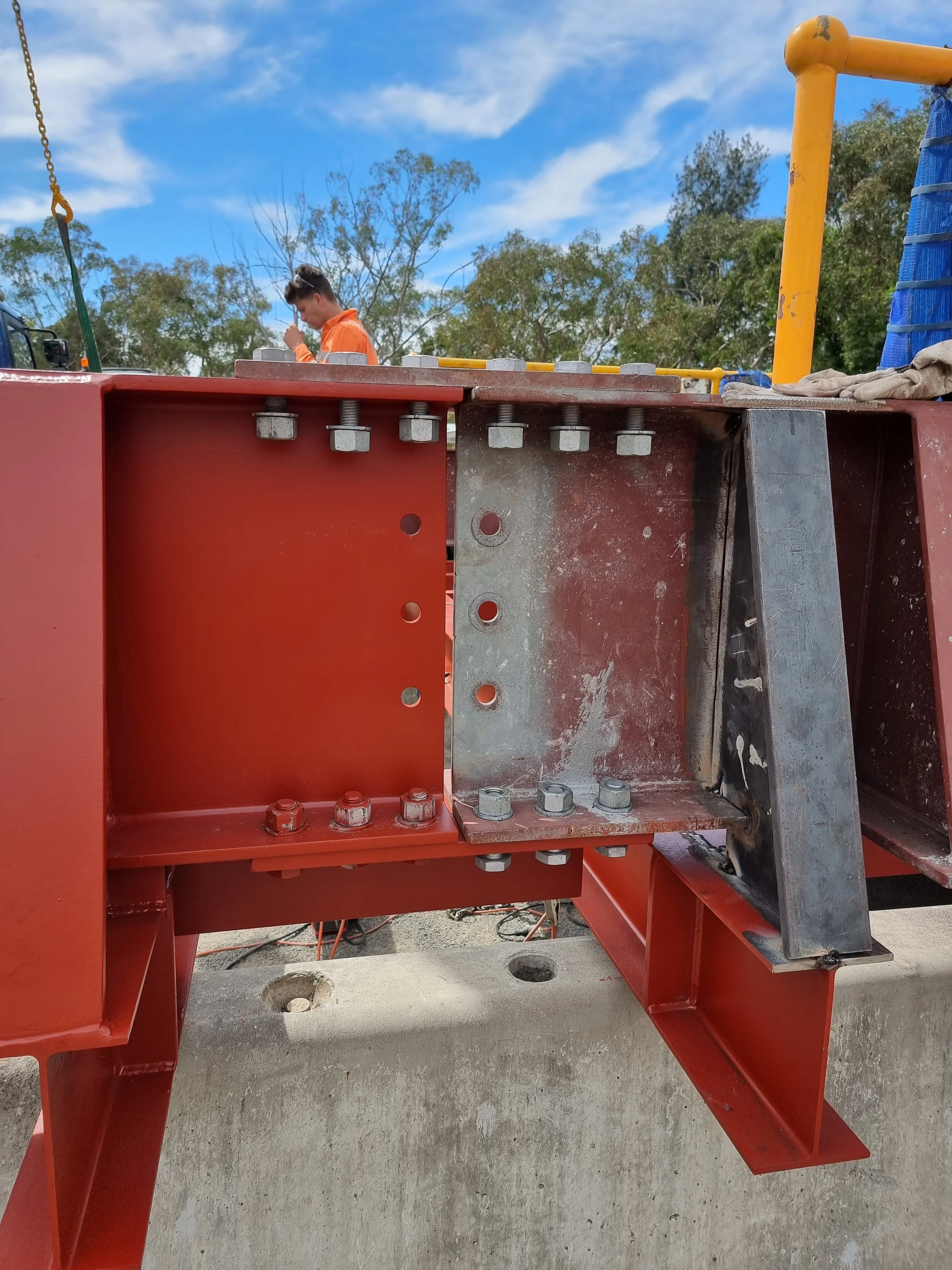 Close-up of a steel structure at a construction site with visible bolts, beams, and a concrete base, with a worker in construction attire in the background under a blue sky with clouds and trees.