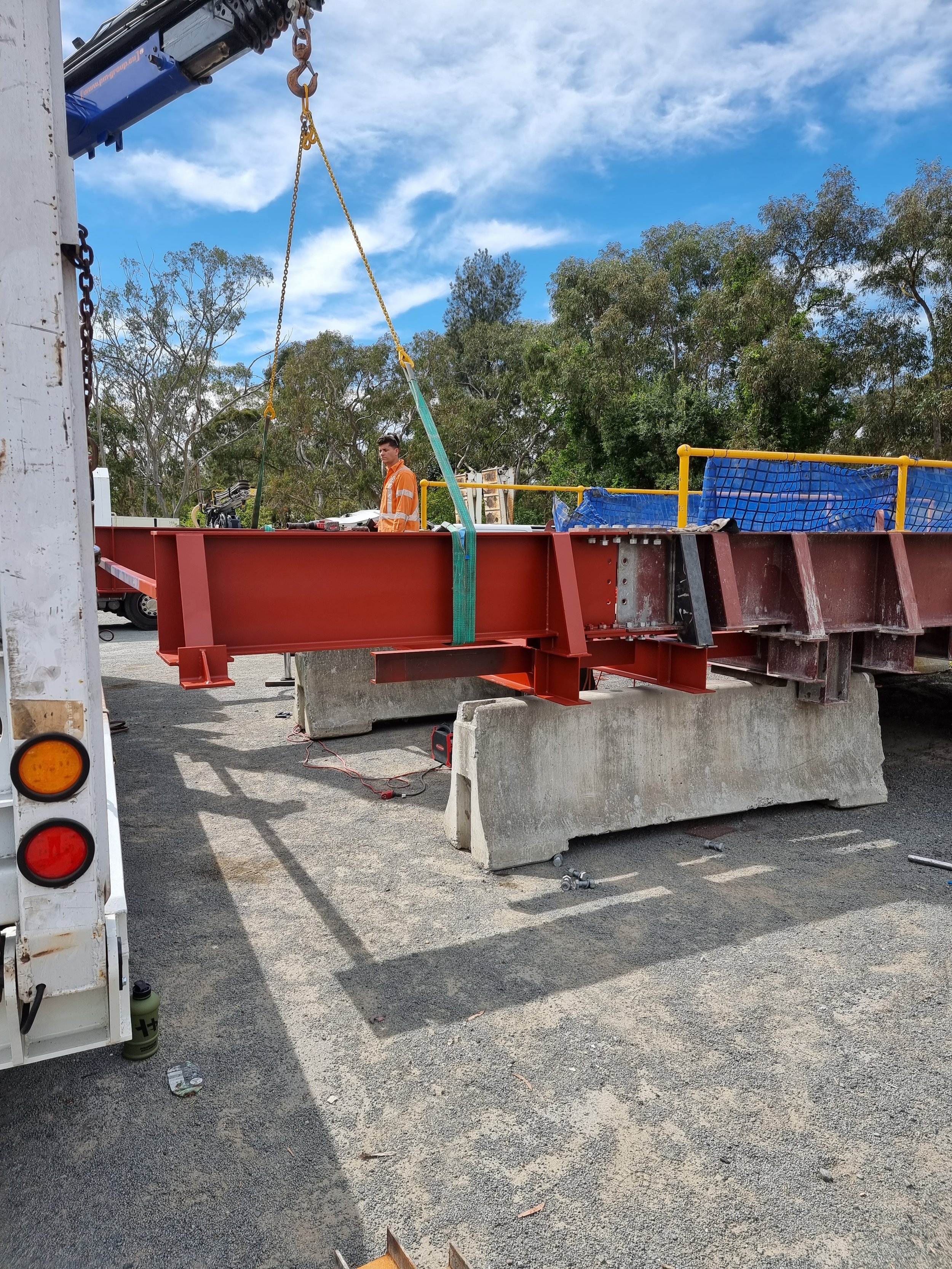 A construction worker standing on a platform next to large concrete blocks, with a crane lifting large steel beams, outdoors with trees and a blue sky.
