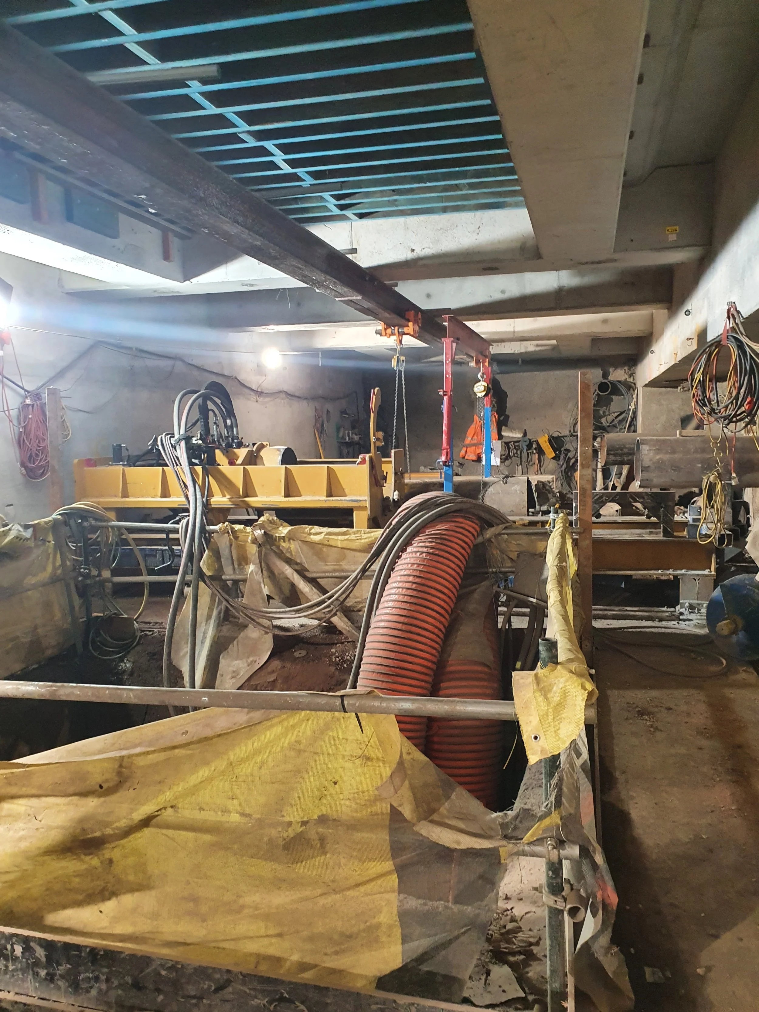 Construction site with orange and gray pipes, yellow safety barriers, and construction equipment under a concrete ceiling.