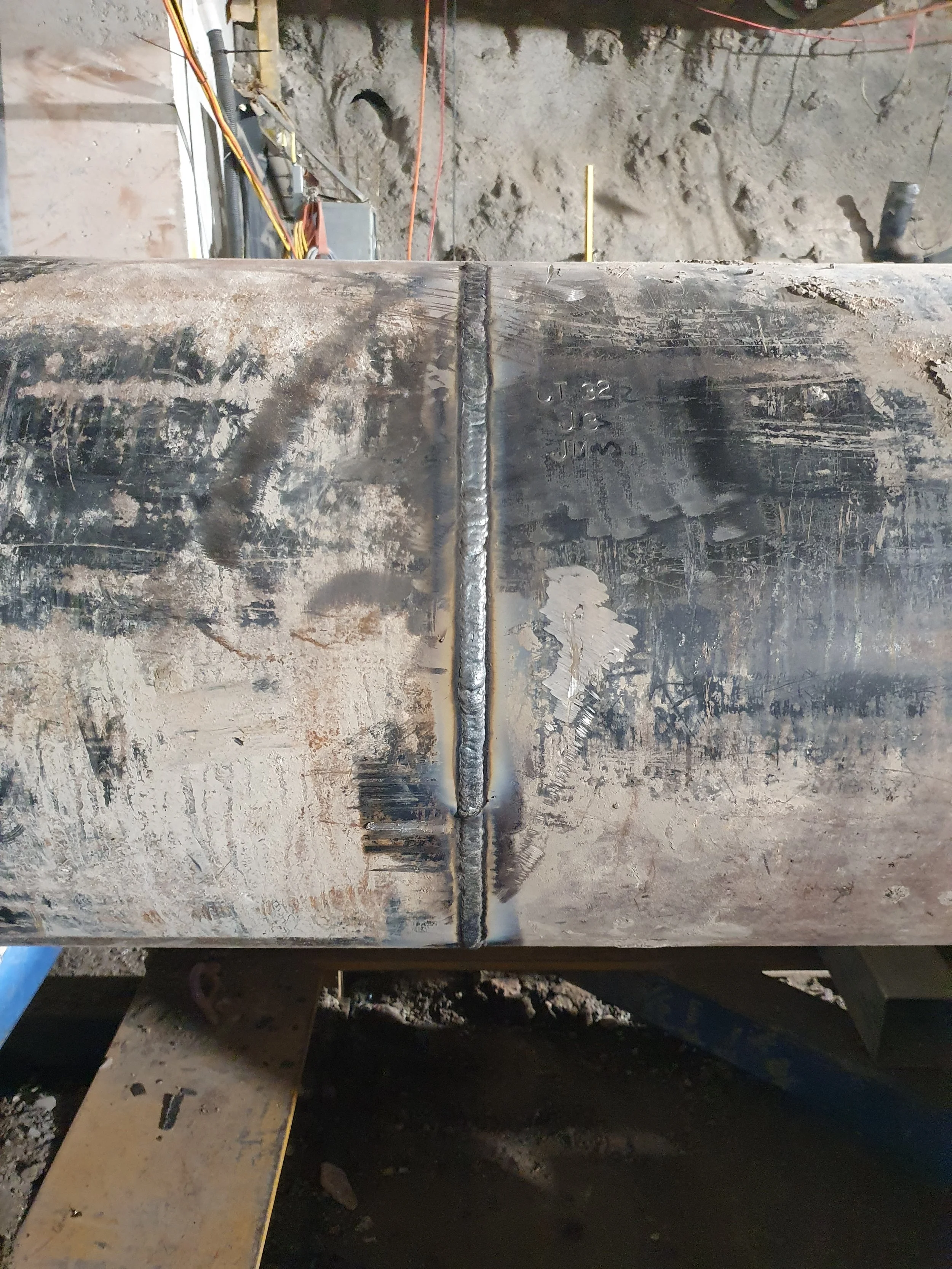 Close-up of a welded metal pipe with a visible weld bead along its length, situated on a construction site with dirt and equipment in the background.