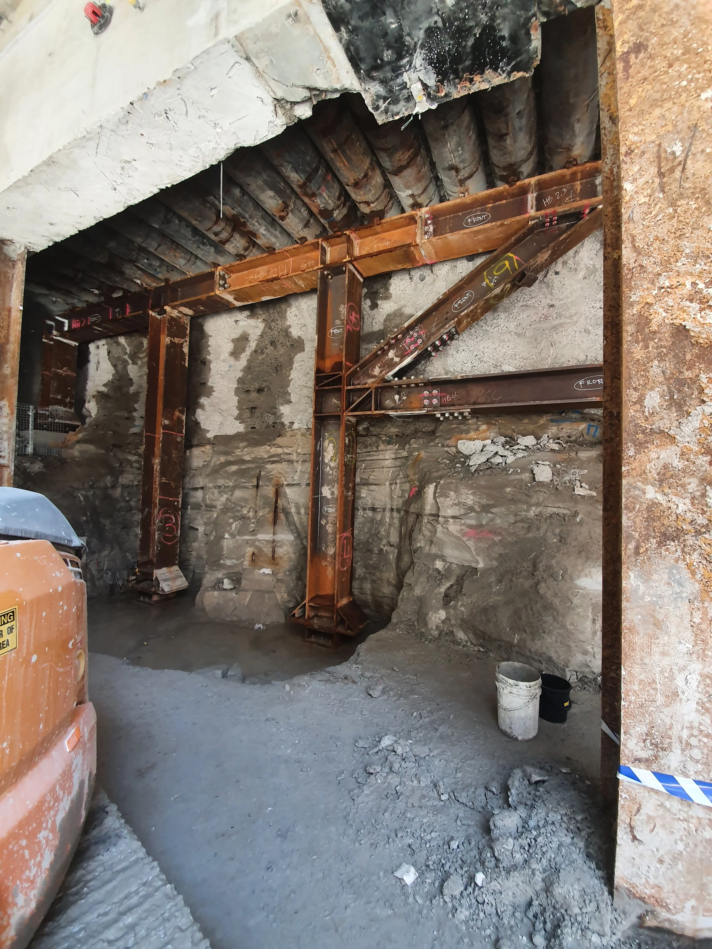 Construction site with exposed steel framework and concrete wall, dirt ground, and construction equipment.