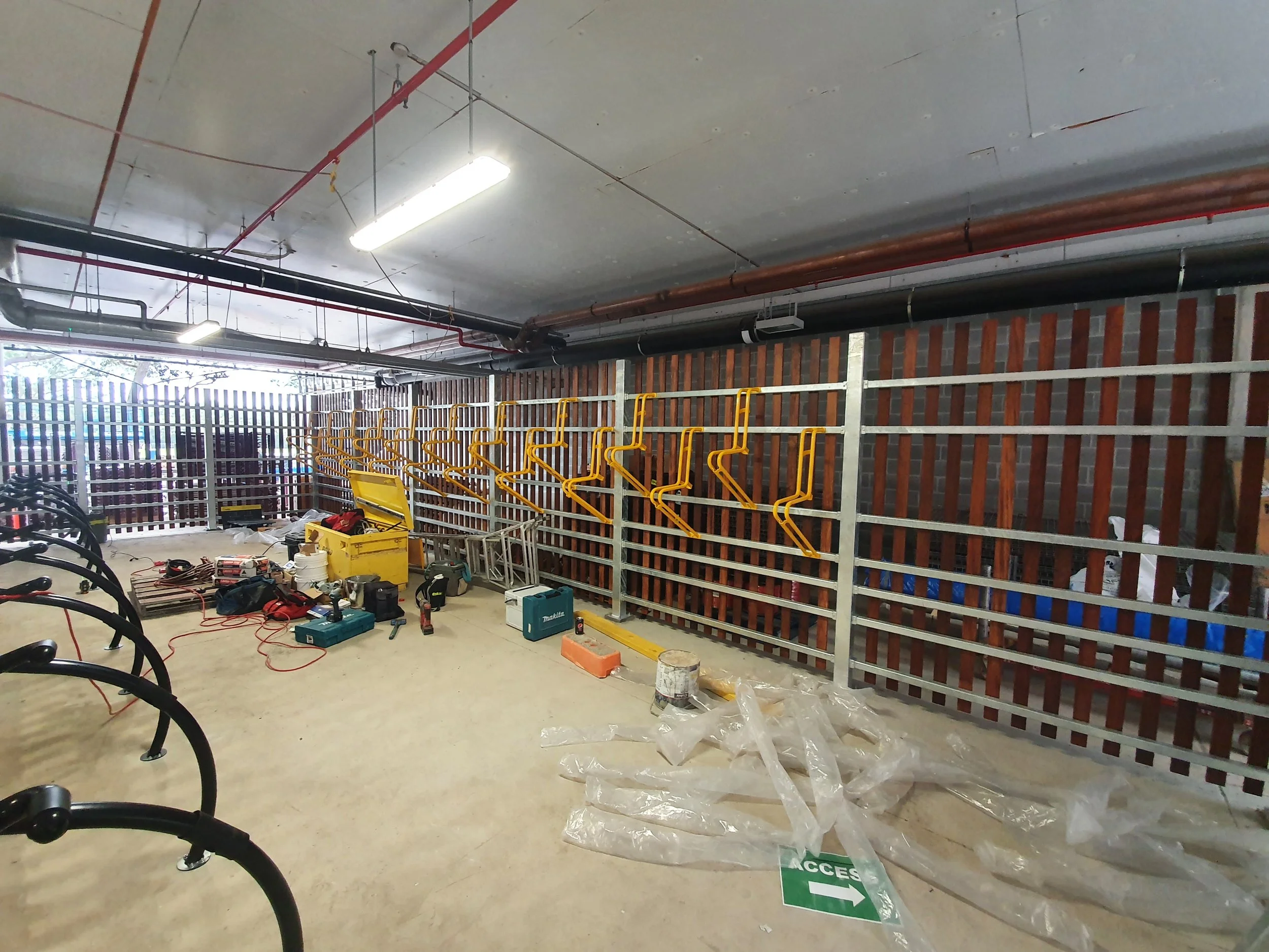 Construction site of a bike parking area with yellow bike racks and tools and materials scattered on the floor. A sign indicates the access point. The background includes a decorative wall with vertical wooden slats.