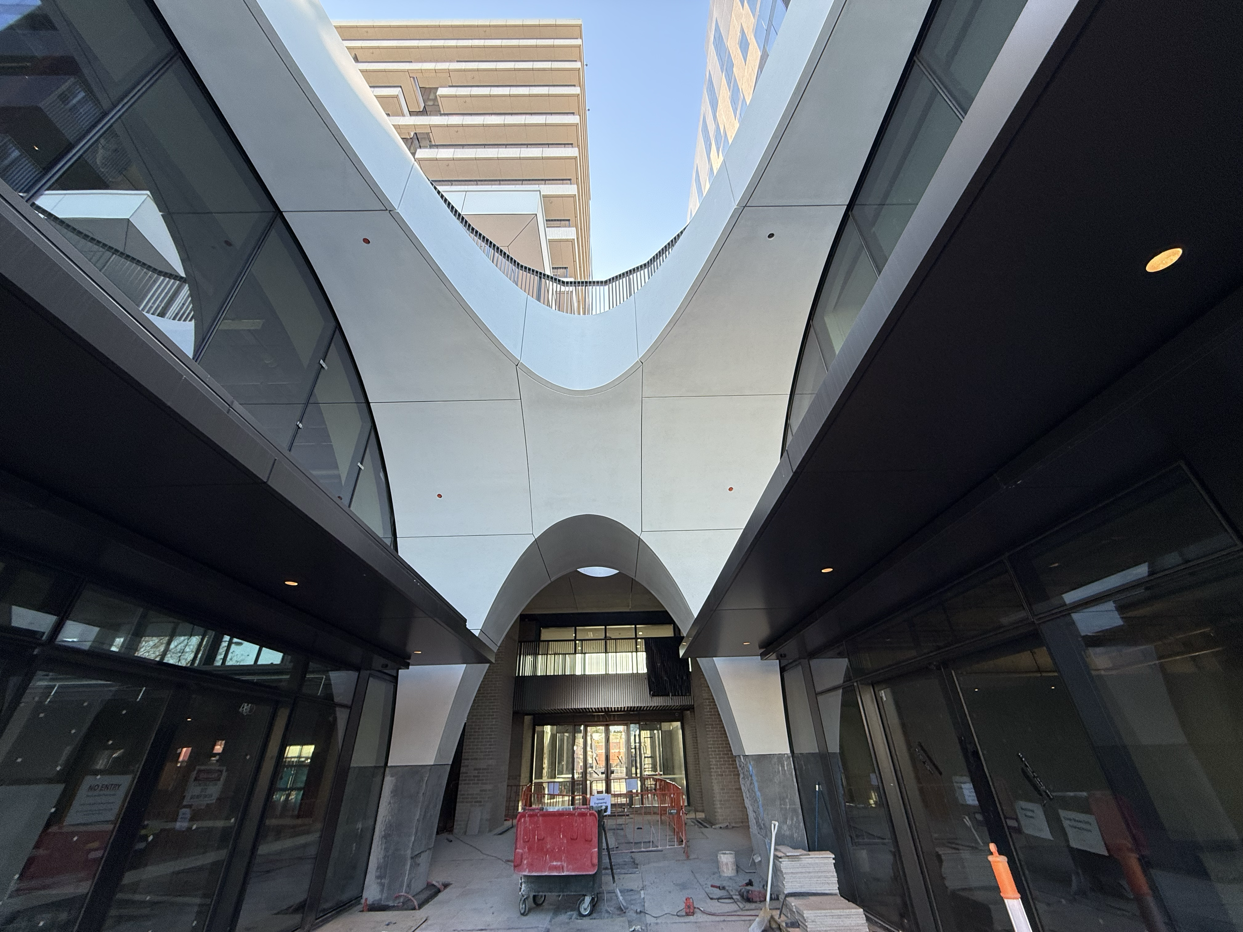 Modern building under construction with an archway and glass windows, construction equipment and barriers in the foreground.