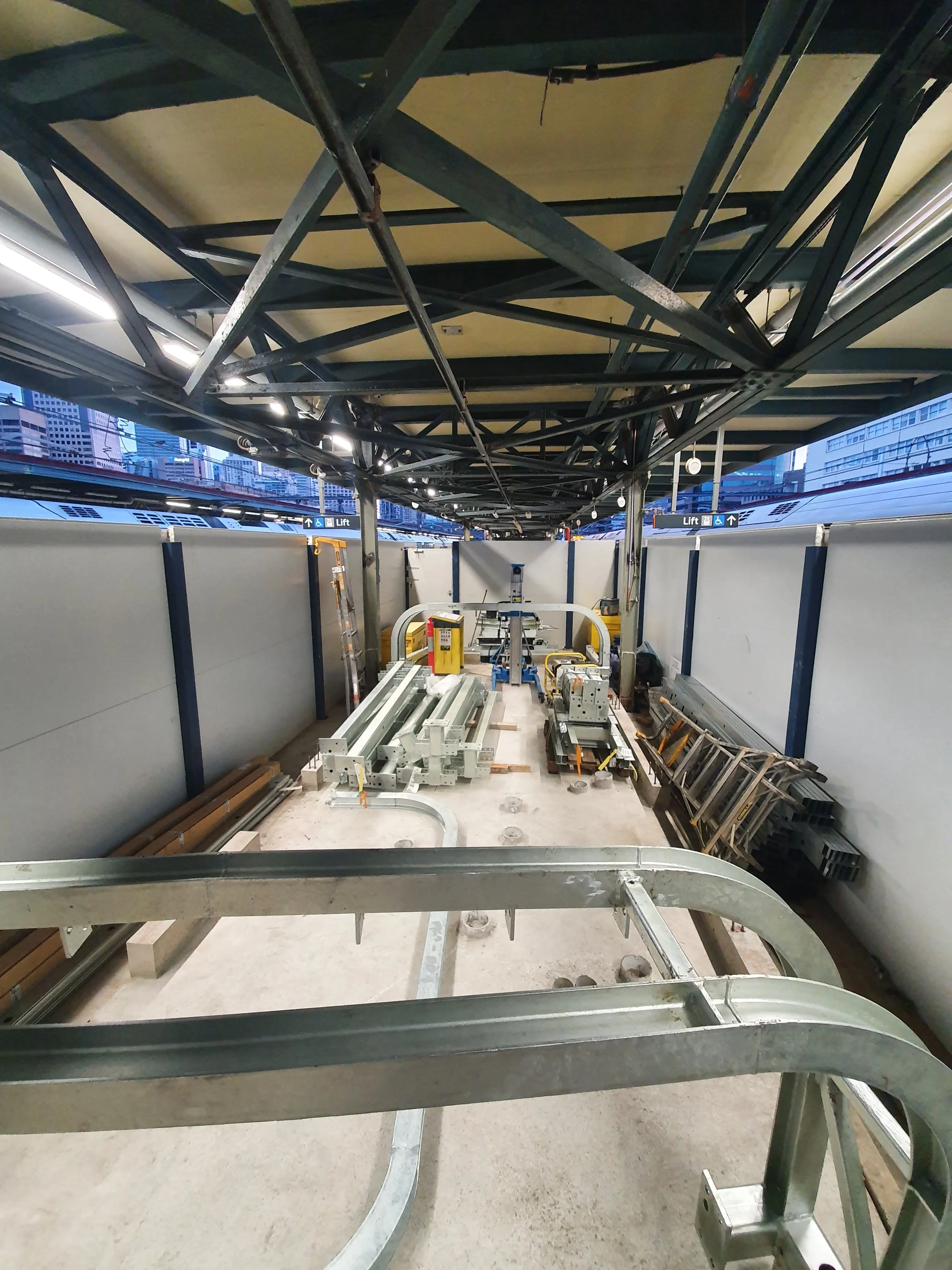 Construction site under a roof, with metal beams, construction materials, ladders, and equipment.