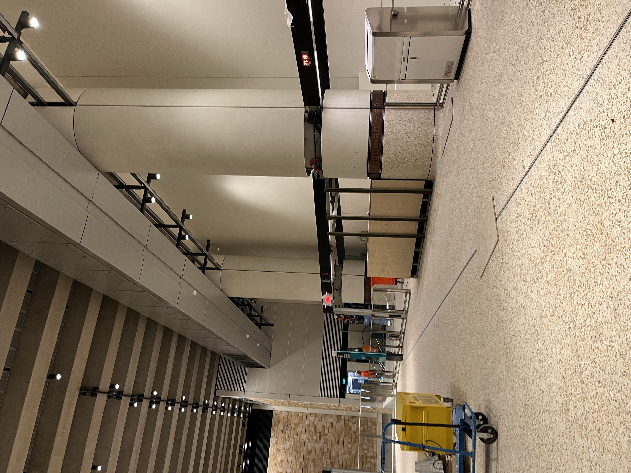 Empty airport or train station lobby with a staircase, cleaning cart, and security checkpoint with scanning machines and orange cones