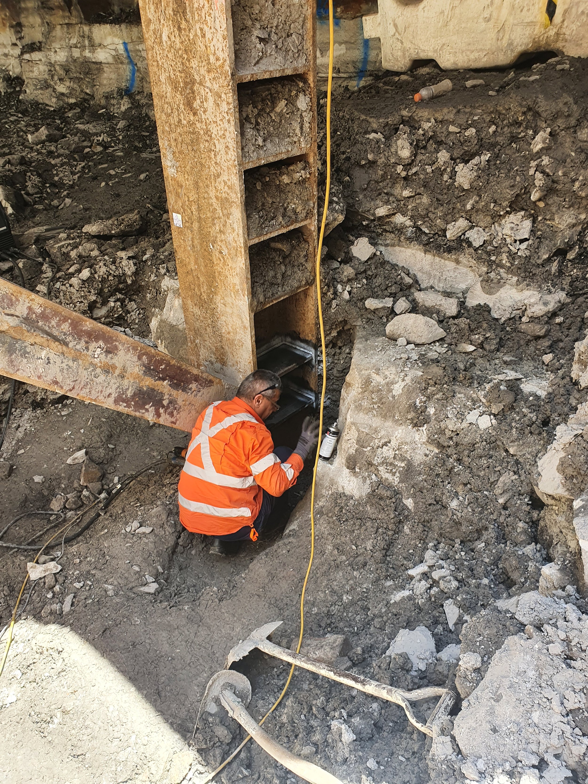 A construction worker in an orange safety jacket and gloves is installing or repairing a drainage or sewer system in a trench. The worker is using a spray can, and there are construction tools and dirt around.